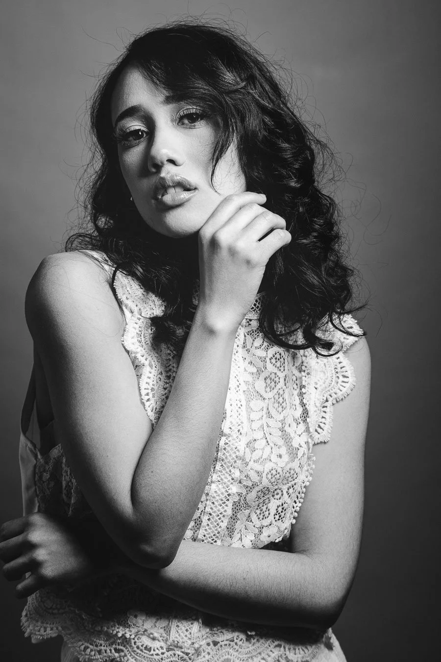 Portrait en noir et blanc d'une femme avec des cheveux bouclés, portant une veste en dentelle, levant la main vers son visage.