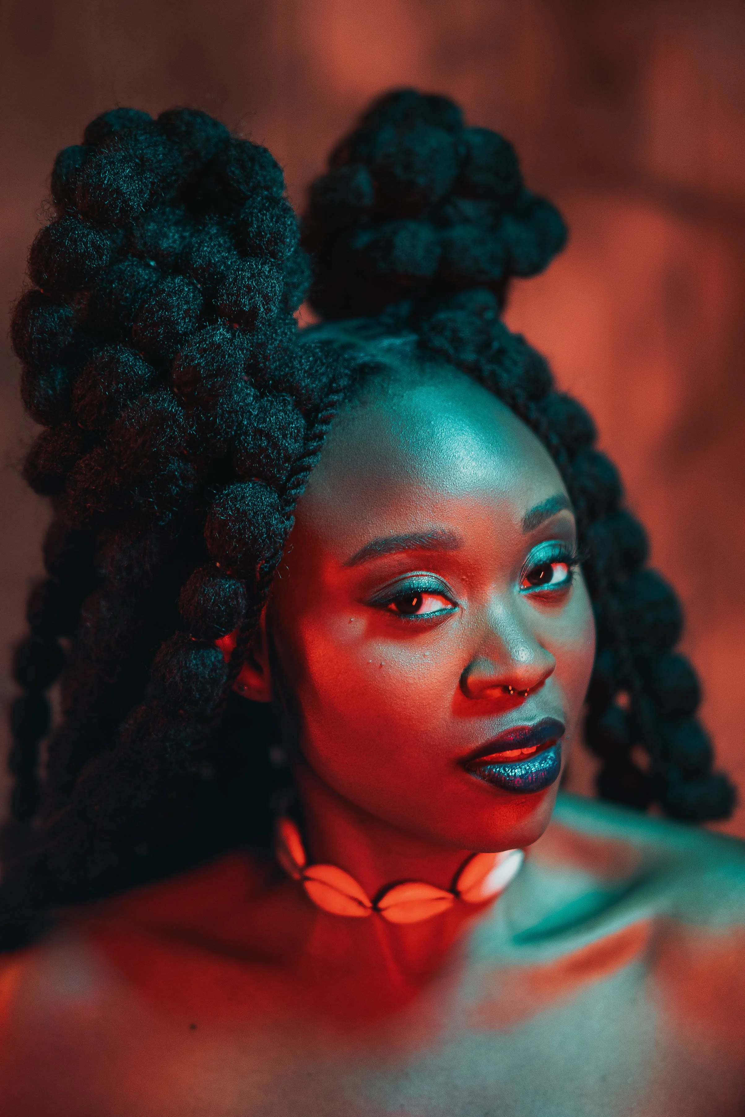 Portrait d'une femme noire avec de grandes tresses bouclées, maquillage sophistiqué, et portant un collier en coquillage. Éclairage coloré rouge et bleu.