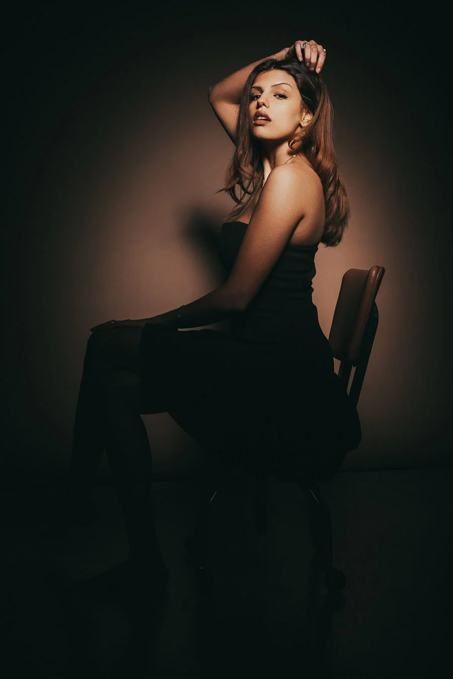 Une femme assise sur une chaise, posant de profil avec une main dans les cheveux, portant une robe noire, dans un éclairage tamisé.