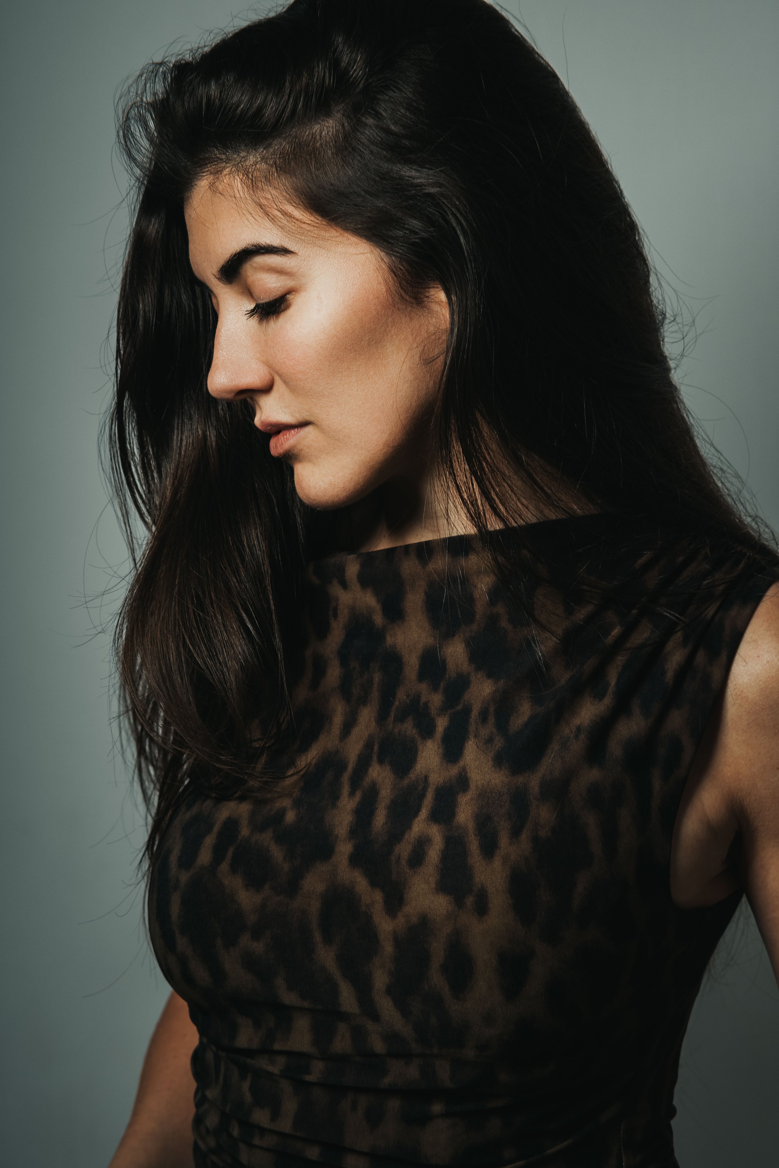 Une femme aux cheveux longs et noirs portant un haut en tissu avec un motif léopard, regardant vers le bas, sur un fond gris.