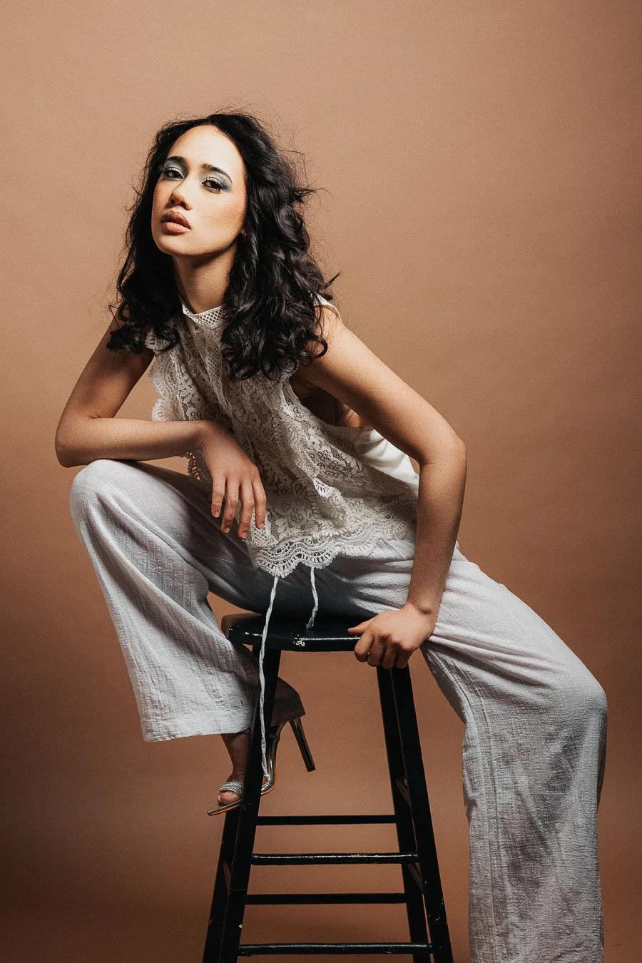 Une femme assise sur un tabouret, portant une tenue blanche en dentelle, contre un fond beige.