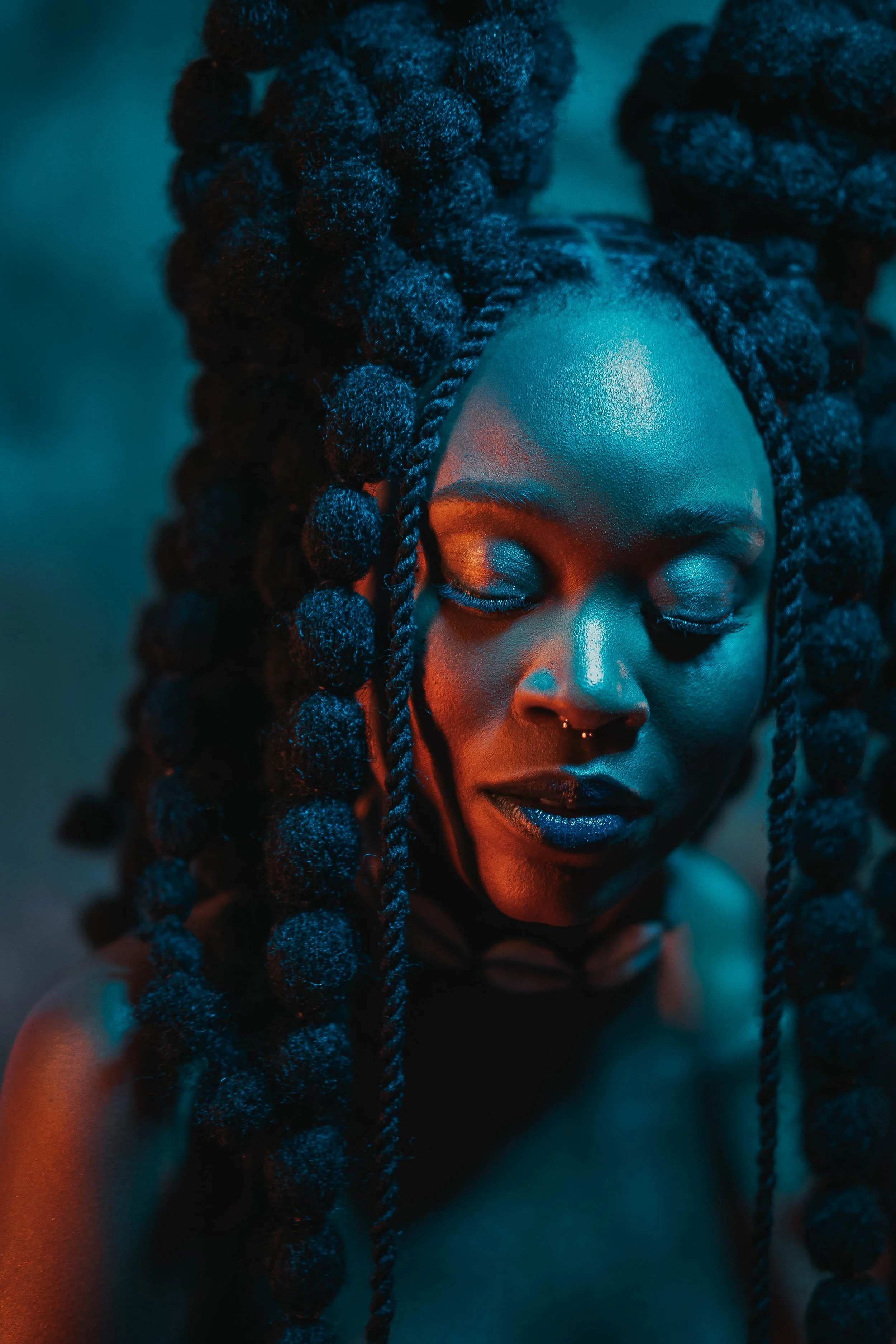 Portrait d'une femme noire aux cheveux tressés en boules, yeux fermés, éclairée par une lumière bleue et orange.