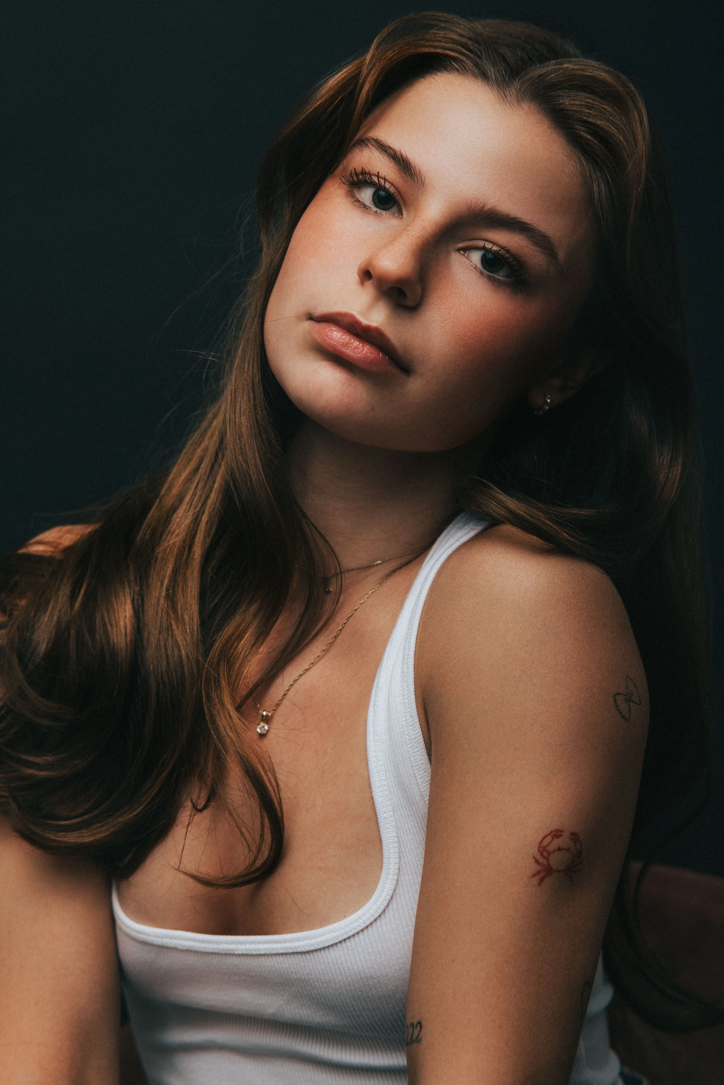Jeune femme aux cheveux longs et bruns, portant un débardeur blanc, avec des tatouages sur le bras, portant un collier avec pendentif, regardant vers l'appareil dans un fond sombre.