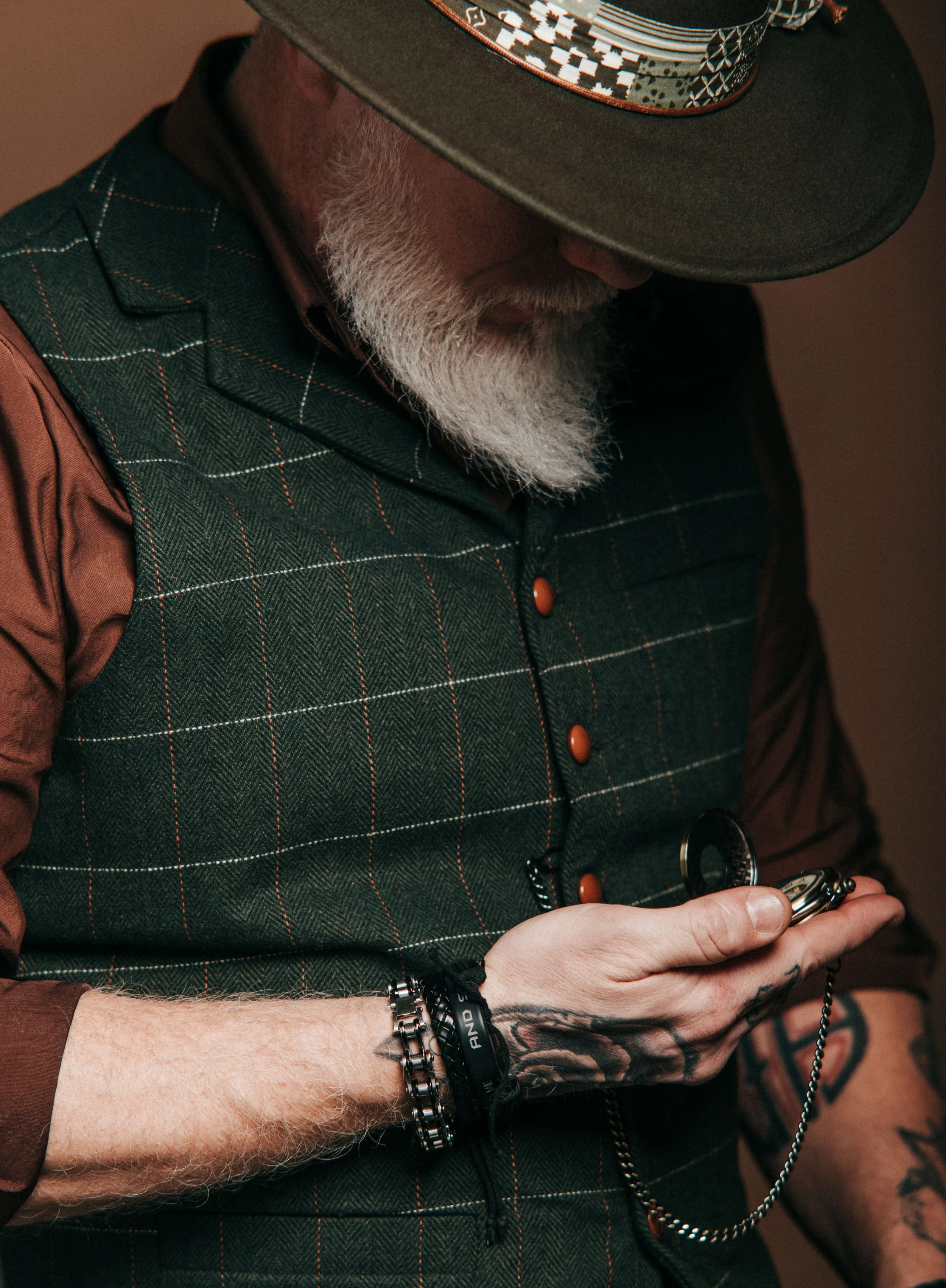Un homme âgé, avec une barbe blanche, portant un chapeau, une veste à carreaux et une chemise marron, regarde une montre à gousset dans sa main droite, avec plusieurs bracelets au poignet gauche.