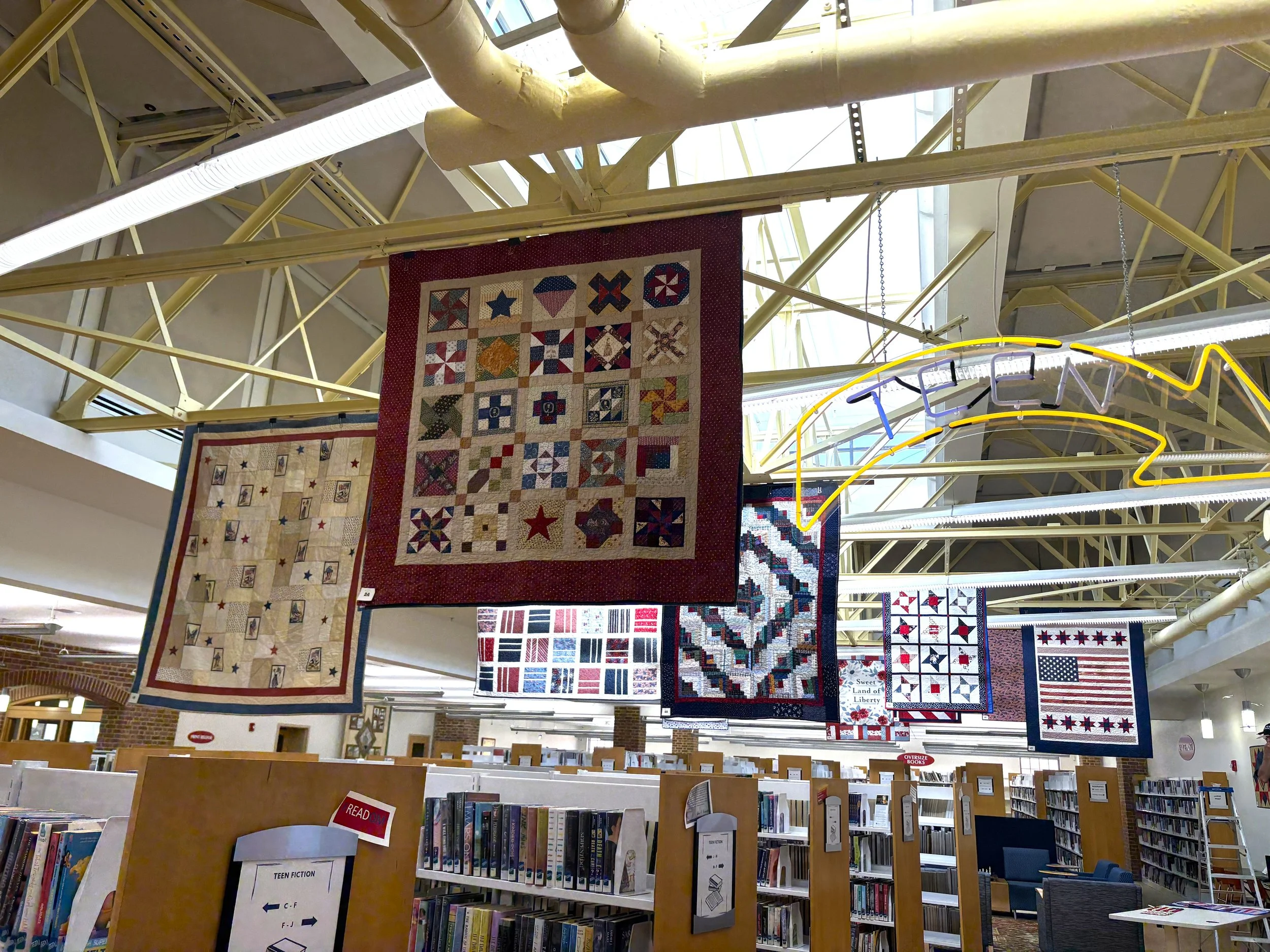 Patriotic quilts hang above book stacks in Talbot County Library during the Bayside Quilters of the Eastern Shore America 250 display.