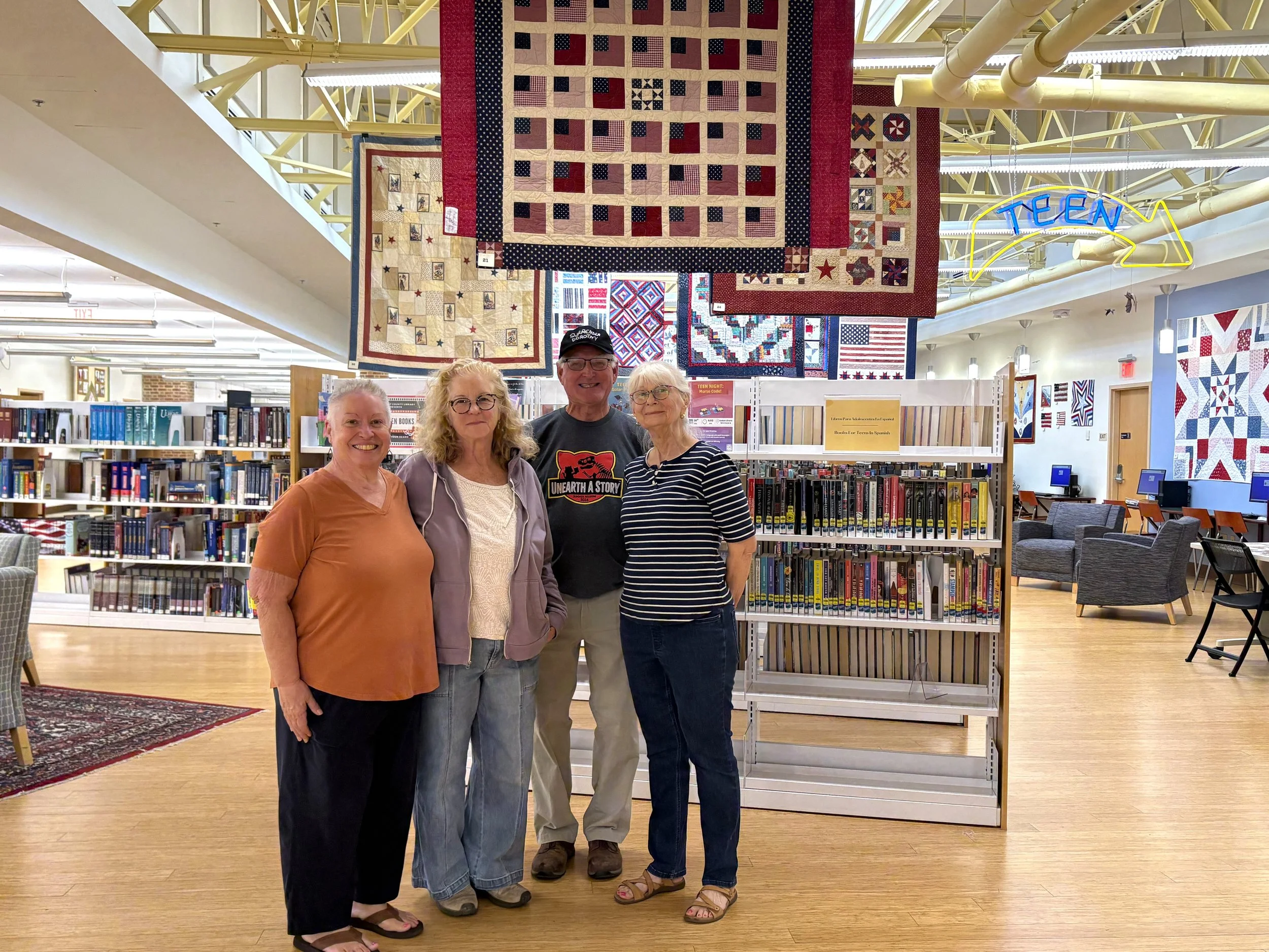 Four Bayside Quilters members stand beneath patriotic quilts at Talbot County Library's America 250 exhibit