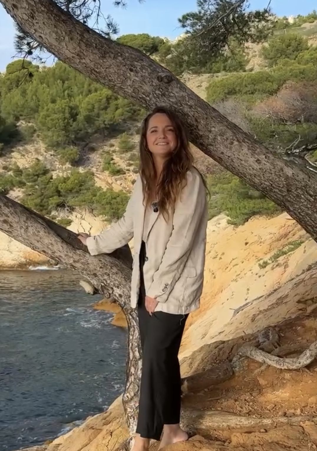 Femme debout sur des rochers près de la mer, tenant une branche d'arbre, paysage naturel avec arbres et collines en arrière-plan, ciel bleu.