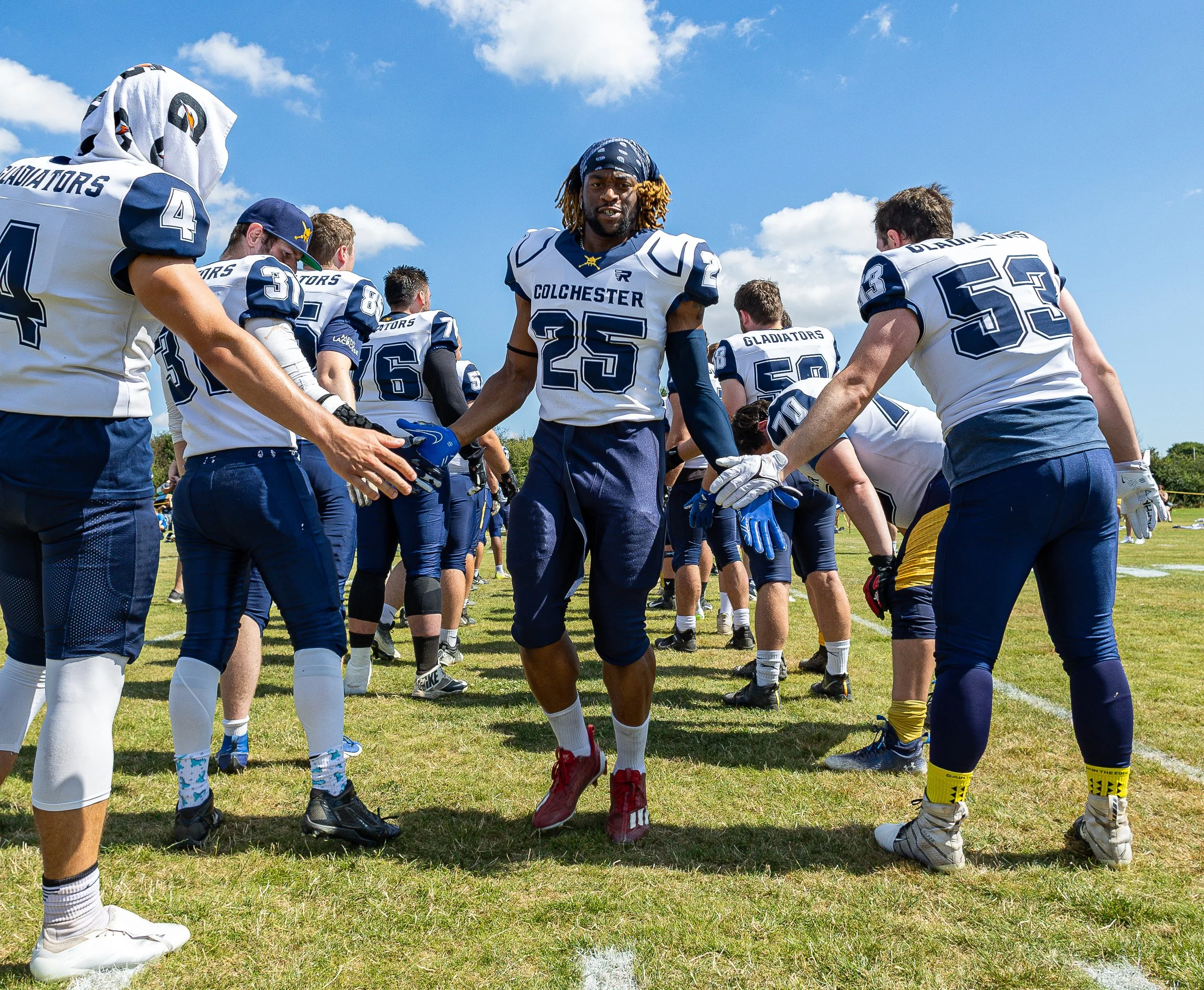 American football players from the Colchester Gladiators team huddling on a grassy field during a sunny day, with a clear blue sky and scattered clouds in the background.