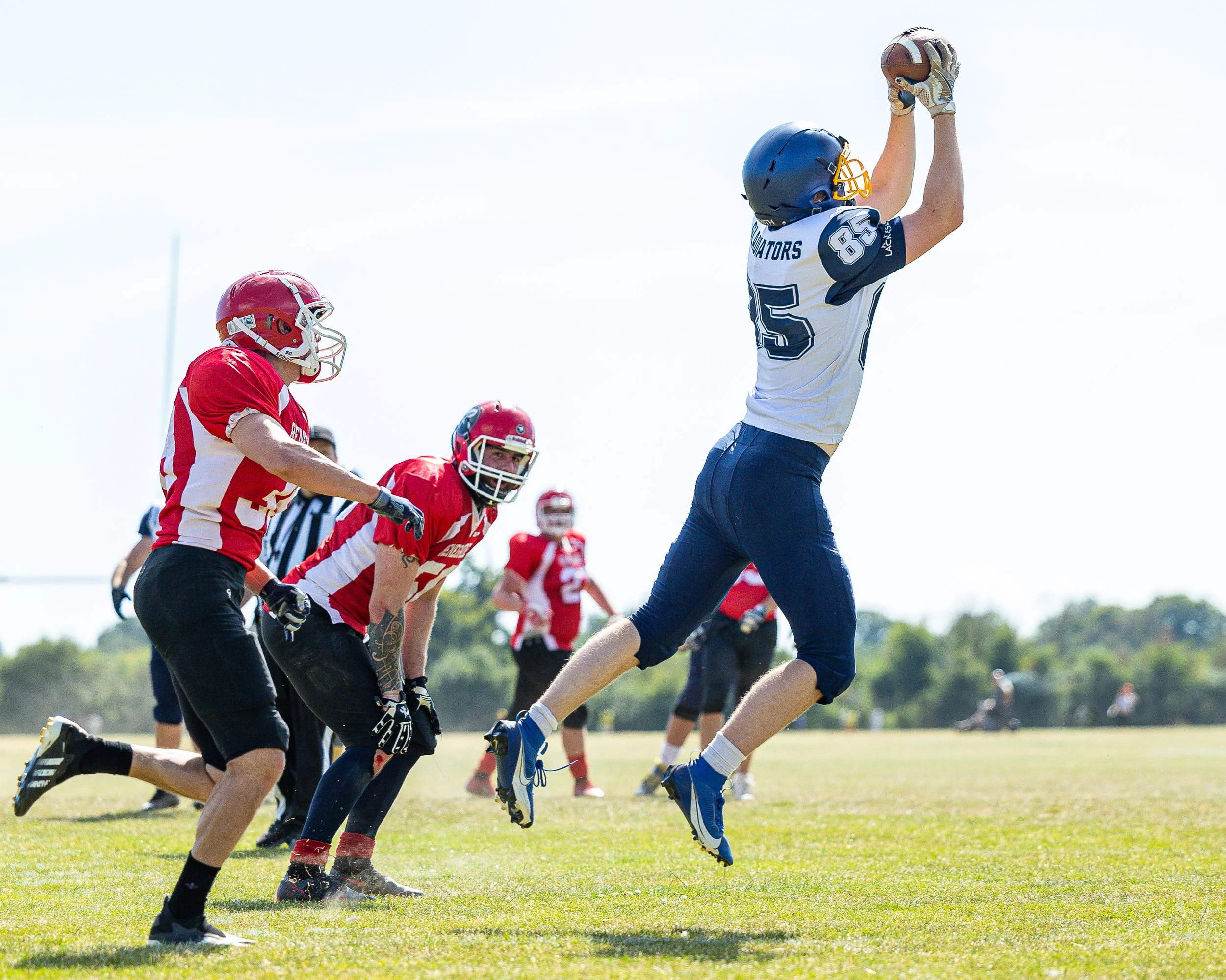 Football players in uniform playing on a grassy field during daytime. One player is jumping to catch the ball, while others are preparing for the play.