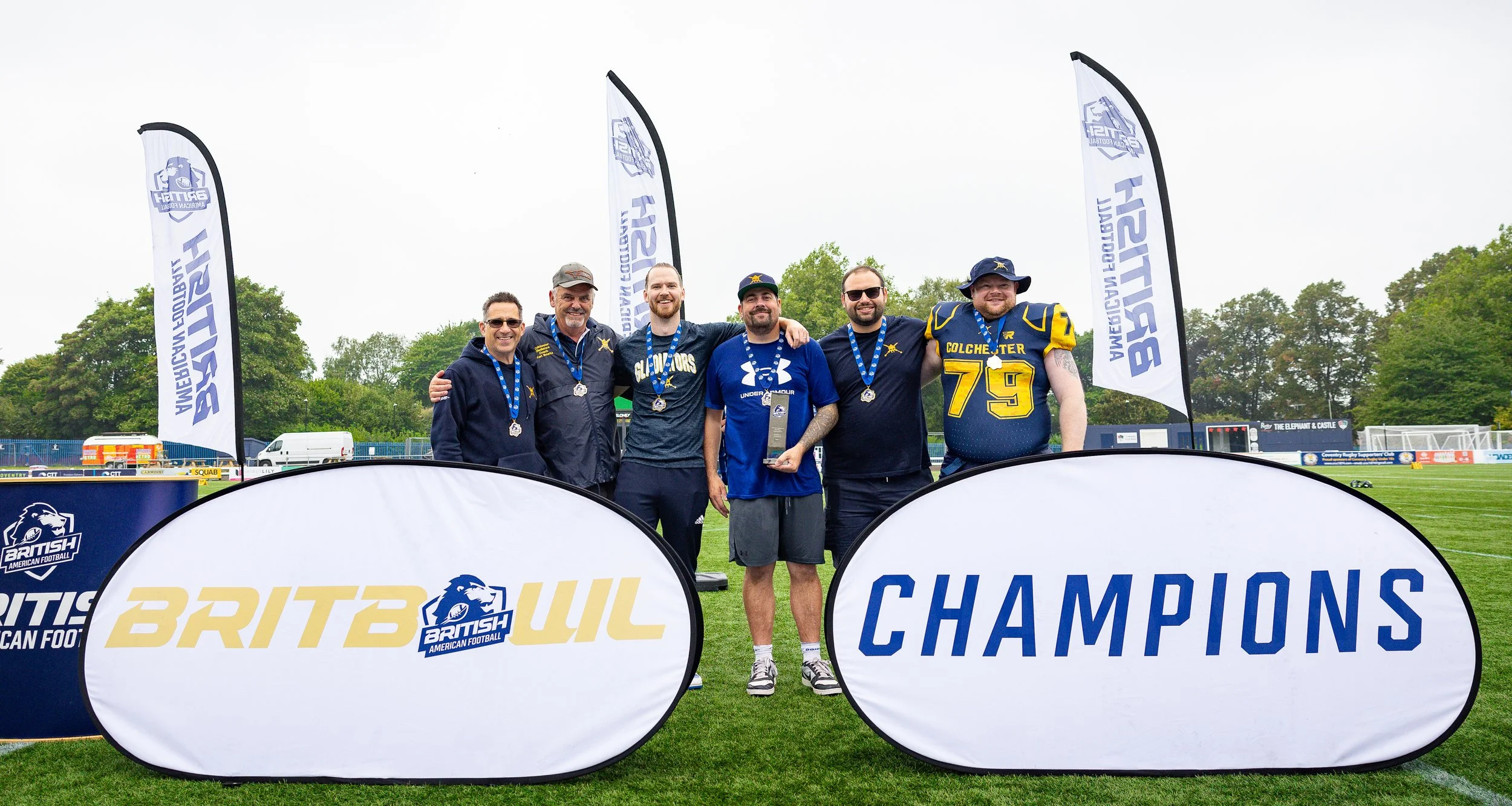 A group of six men standing on a football field, celebrating their victory with medals and a trophy, amid banners reading 'BRITISH AMERICAN FOOTBALL' and 'CHAMPIONS.' The men are smiling, some with arms around each other, dressed in sportswear and casual clothing.