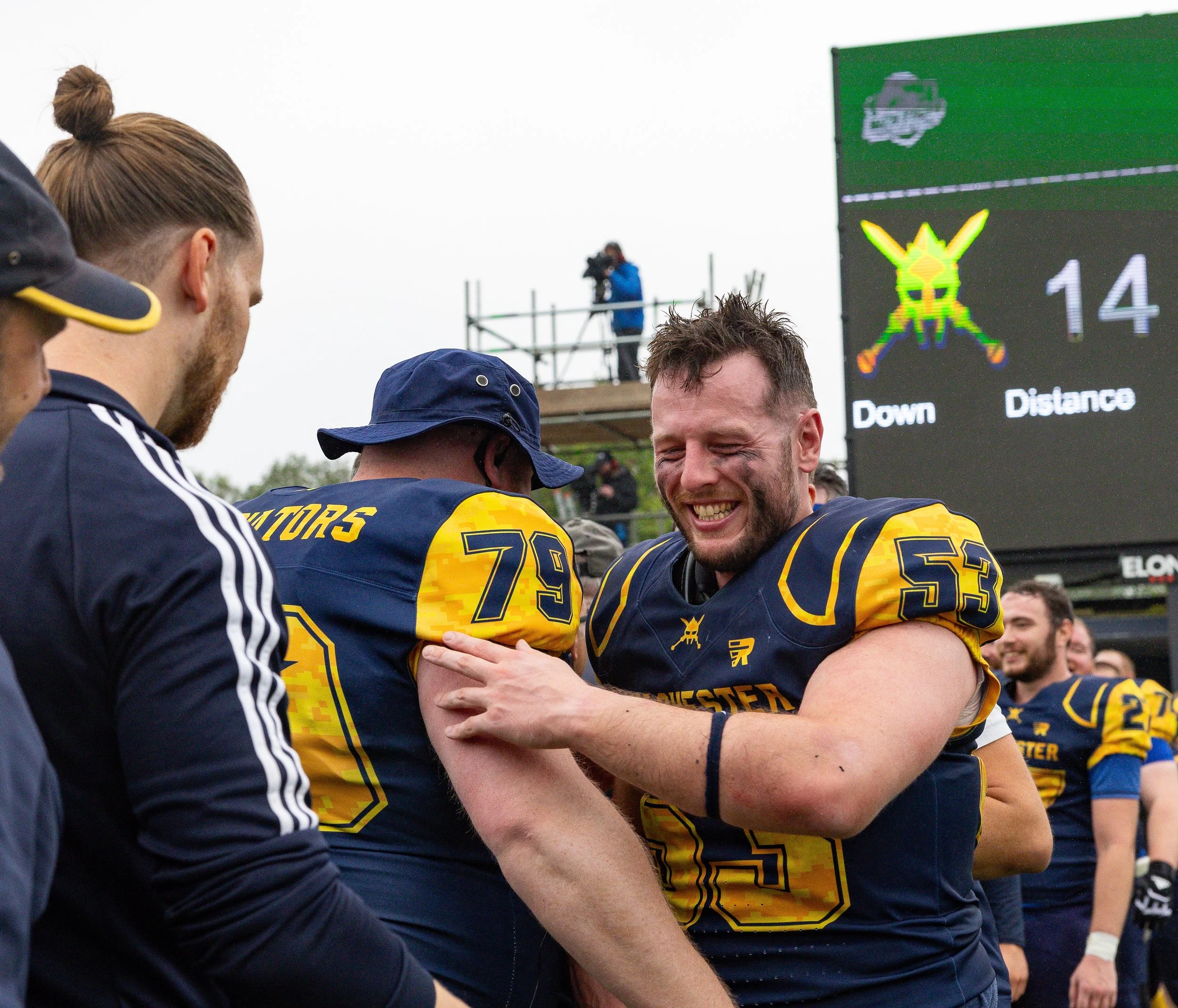 A group of football players in yellow and navy uniforms celebrating on a field, with a large digital scoreboard displaying a bug logo and the number 14 in the background.