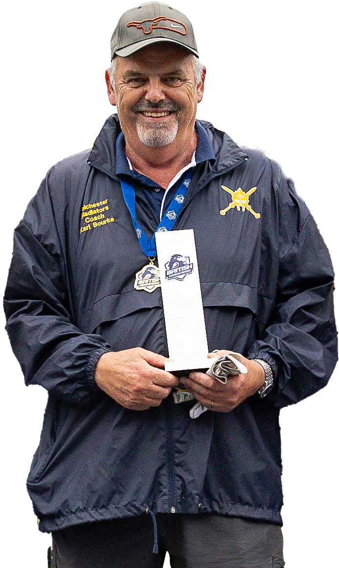 A middle-aged man wearing a gray cap, a dark jacket with a yellow emblem, and a blue lanyard with a medal around his neck, holding a trophy and a golf glove, smiling at a sports event or award ceremony.