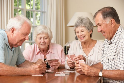 Four elderly people playing cards at a table in a bright living room.