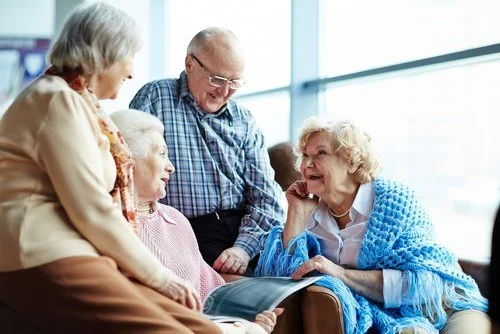 Four elderly people, three women and one man, gathered together, smiling and chatting in a bright room with large windows.