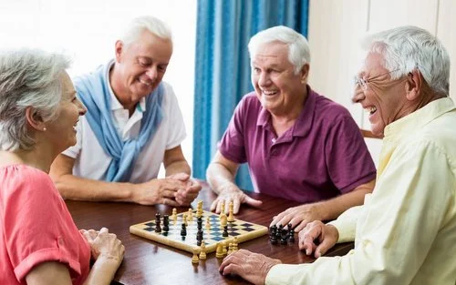 Four elderly people playing chess together, smiling and enjoying each other's company at a table.
