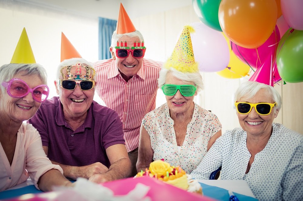 Celebrate a birthday with elderly friends wearing colorful party hats, funny glasses, and balloons.