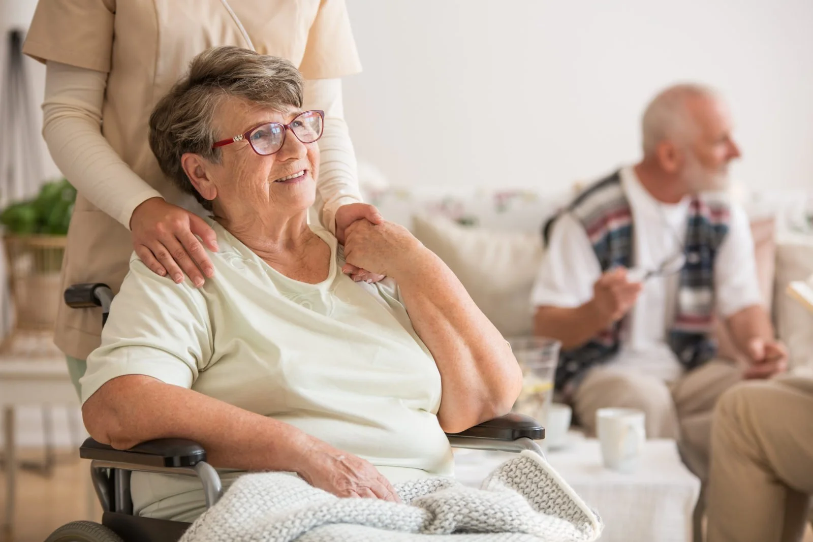 An elderly woman in a wheelchair smiling, with her caregiver standing behind her, supporting her shoulder. Two elderly men are sitting in the background, one holding a spoon and the other with a mug in front of him.