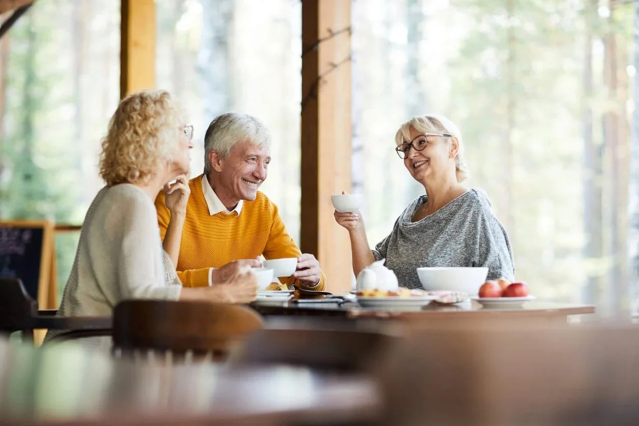 Three elderly people enjoying tea or coffee at a wooden table in a bright, airy room with large windows showing a forested background. They are smiling and engaging in conversation.