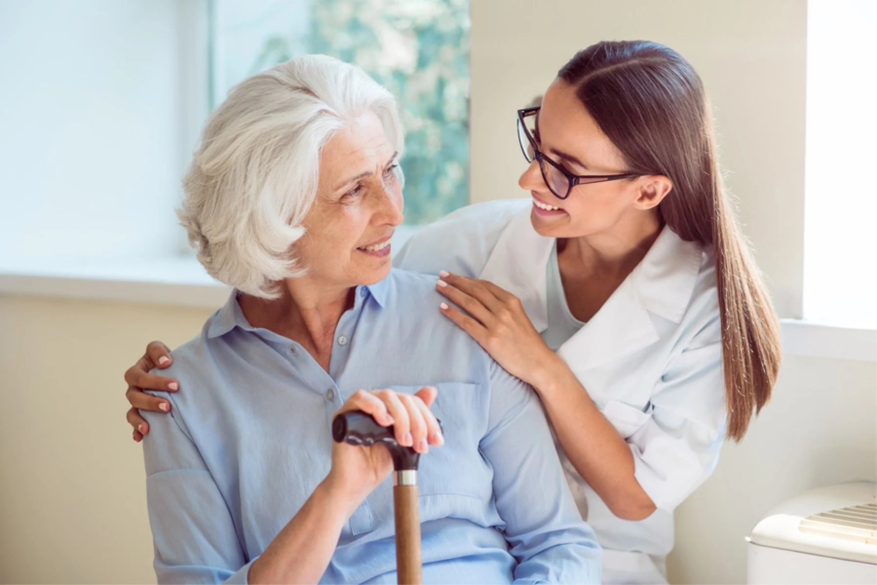 A healthcare worker smiling at an elderly woman holding a cane and sitting on a couch, with the healthcare worker placing a hand on the woman's shoulder.