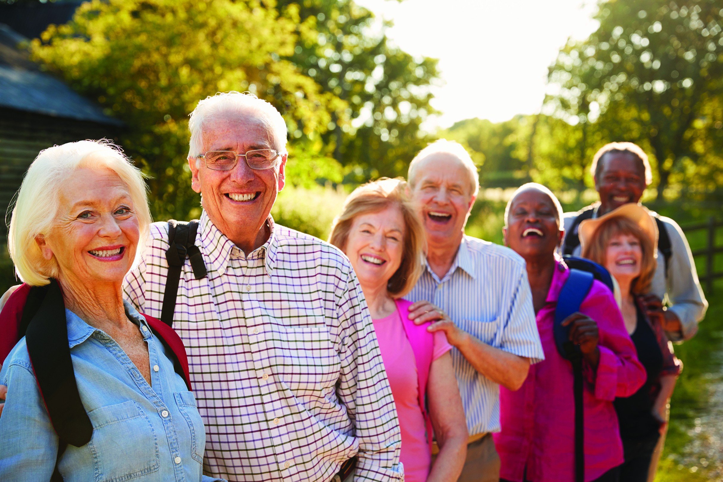 Group of smiling seniors outdoors in a park during daytime, standing in a line with trees and sunlight in the background.