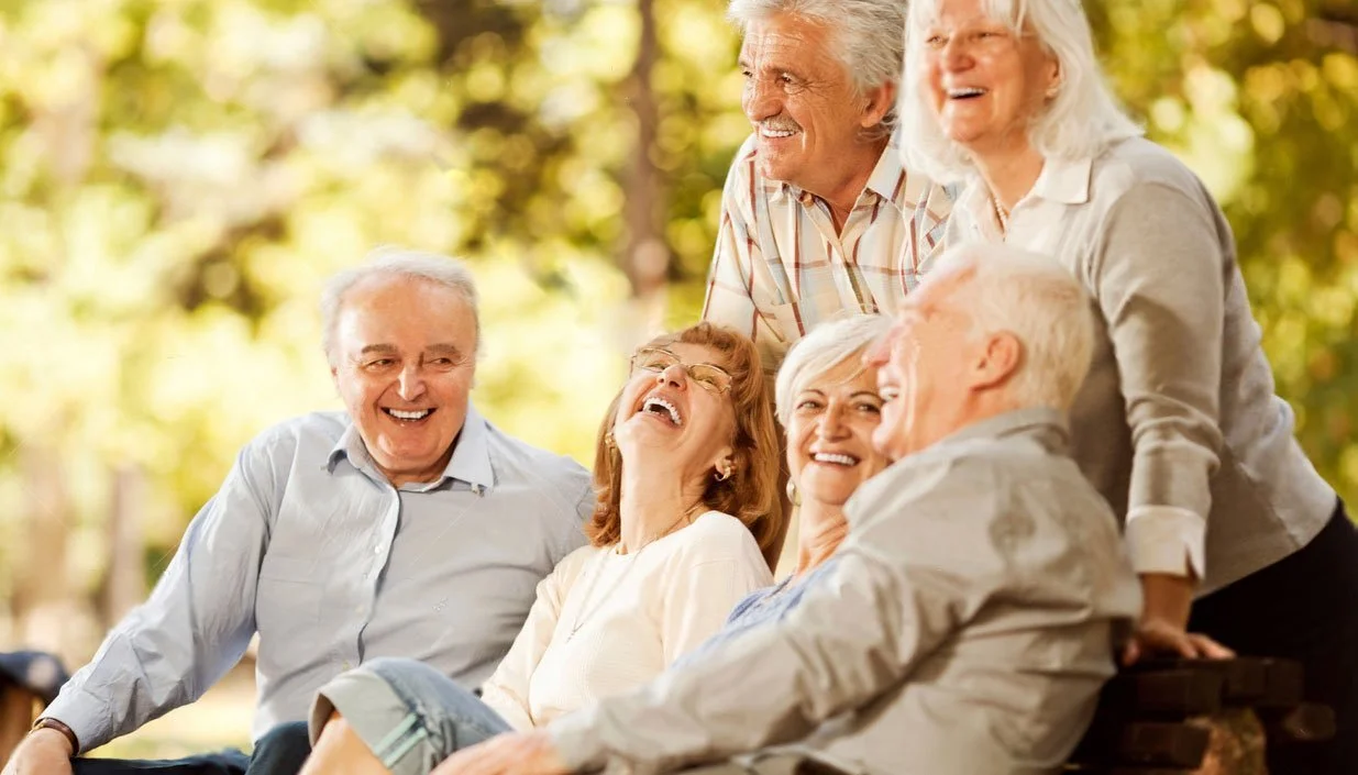 Six elderly people laughing and enjoying each other's company outdoors on a sunny day.