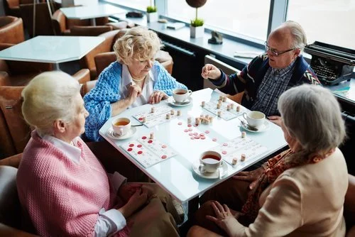 Four elderly people playing a game of checkers at a cafe table, with cups of tea or coffee.