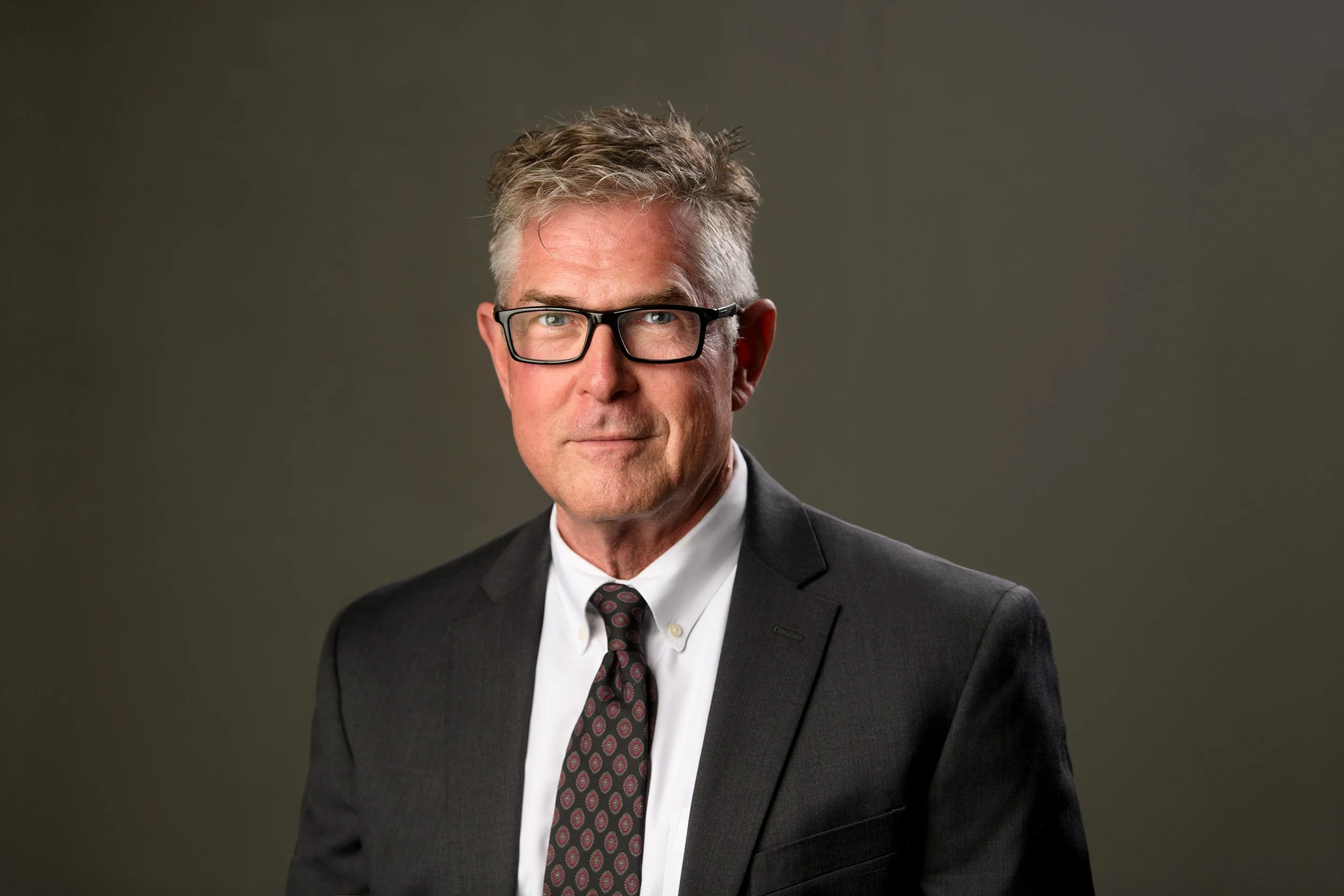 A middle-aged man with gray hair, glasses, wearing a dark suit, white shirt, and patterned tie, against a plain gray background.