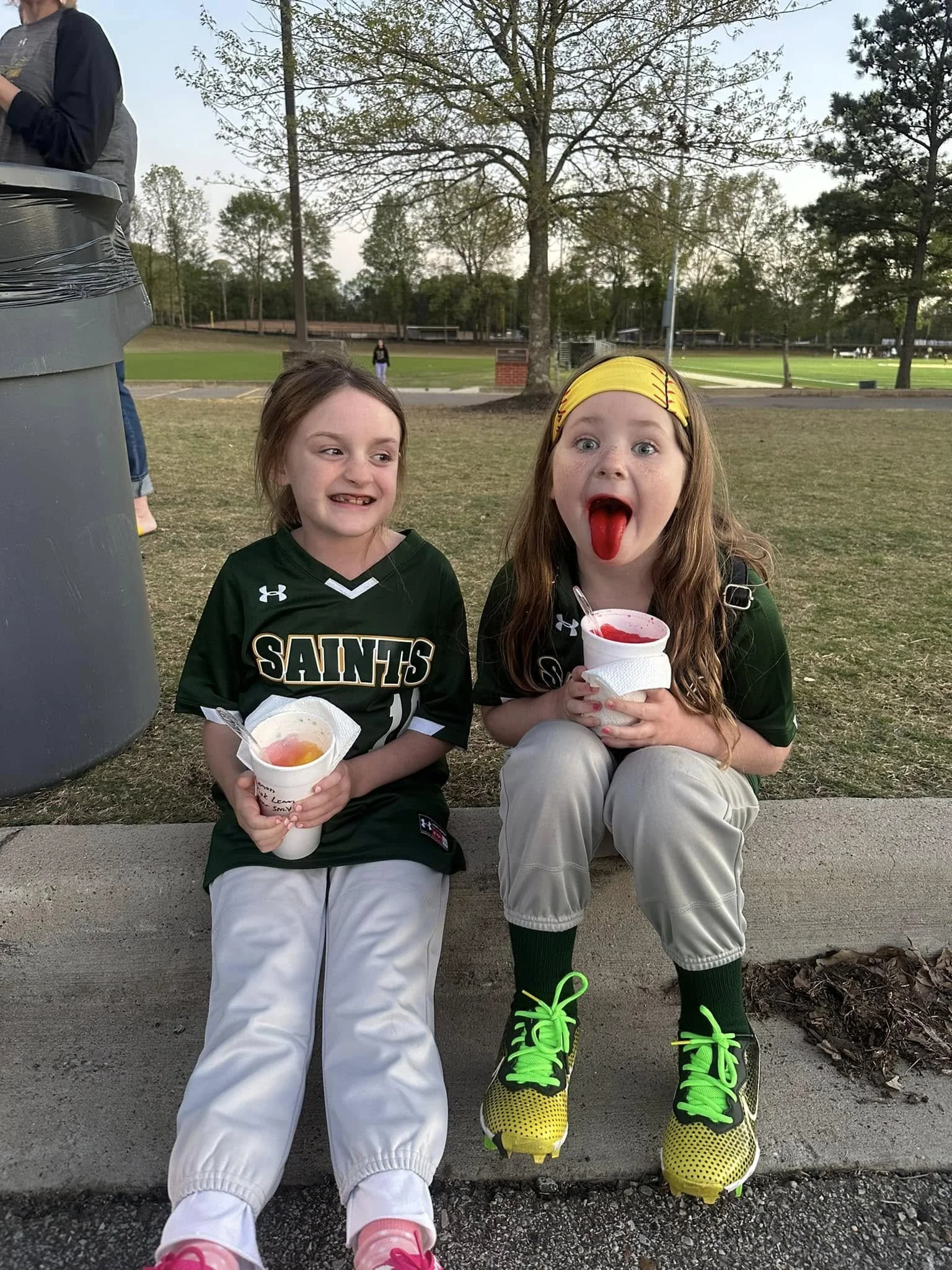 Two young girls sitting on a curb outdoors, enjoying snow cones. One girl is smiling, the other girl is licking her snow cone with her tongue out. They are wearing sports jerseys, and the girl on the right is wearing bright yellow and green sneakers and a yellow headband. Trees and open fields are visible in the background.