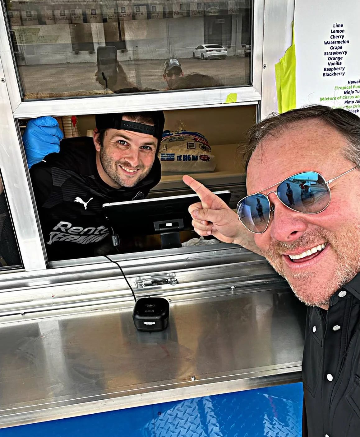 Two people at a food truck window, one inside wearing a cap and gloves, and the other outside pointing and smiling.