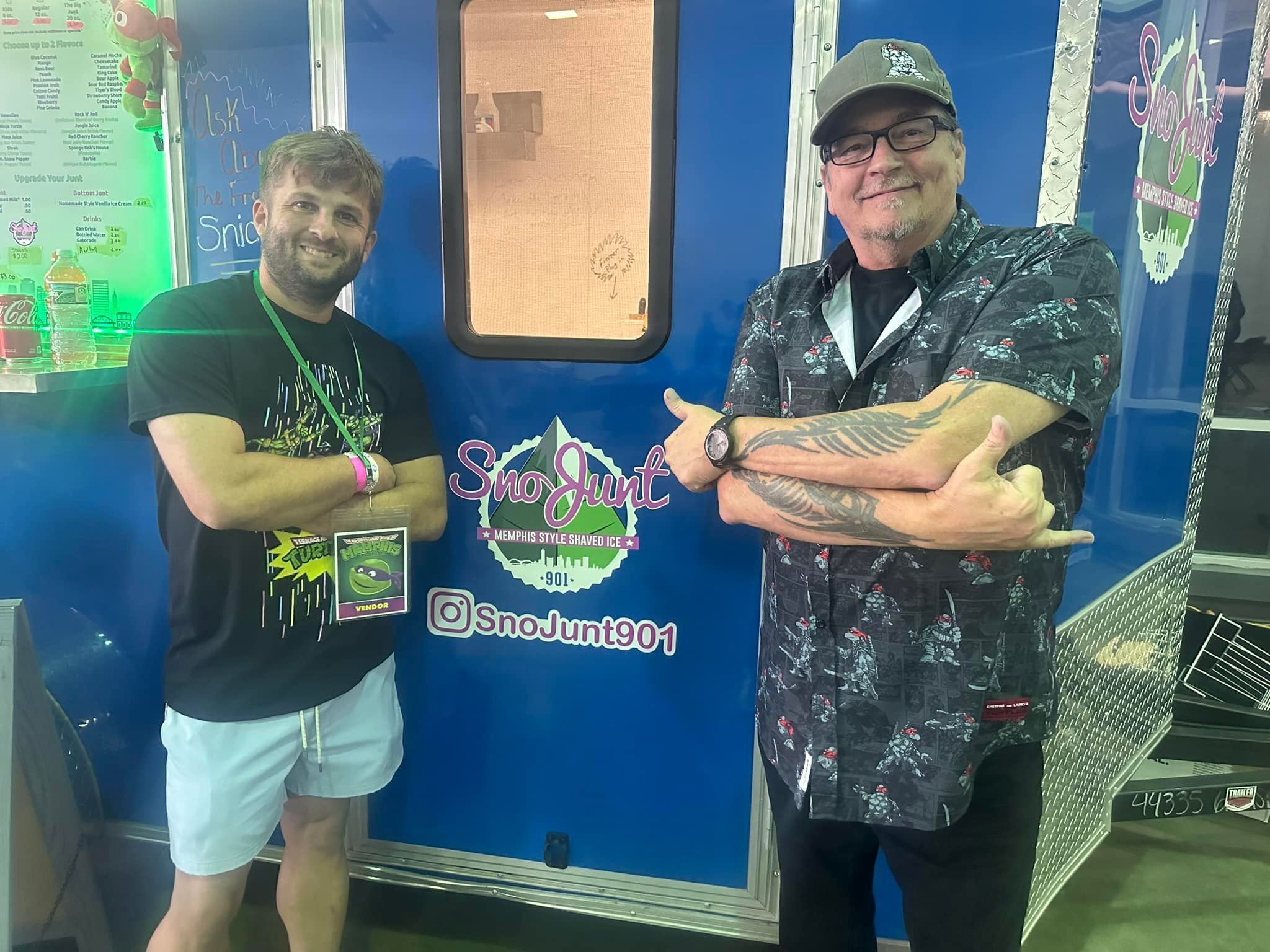Two men posing in front of a blue food truck with the logo "SnoJunt 901," offering Memphis-style shaved ice. One man is wearing a black T-shirt and shorts, and the other is wearing a patterned shirt and cap, both smiling.