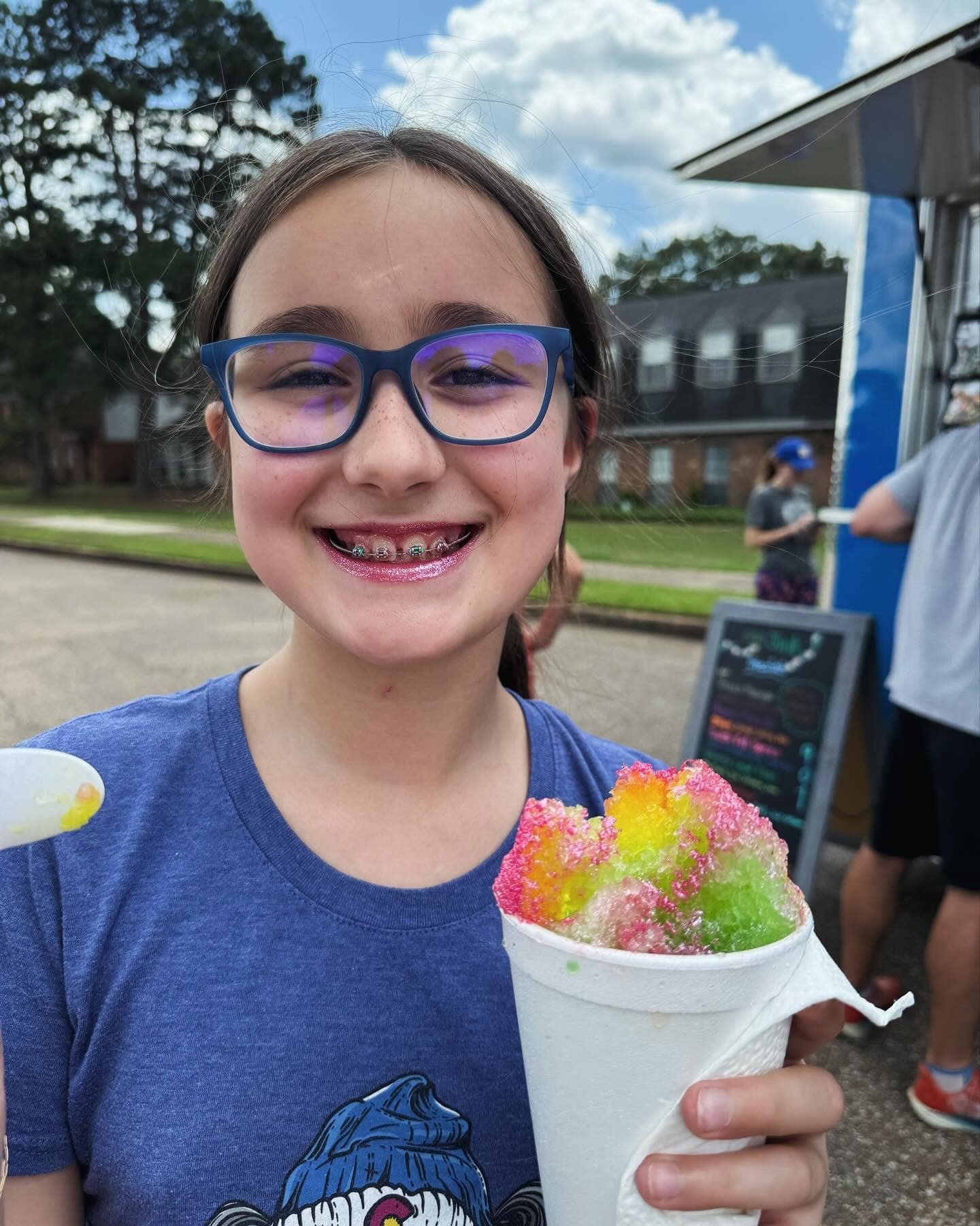 Girl with glasses and braces holding a colorful snow cone outside, wearing blue shirt, with trees and food truck in the background.