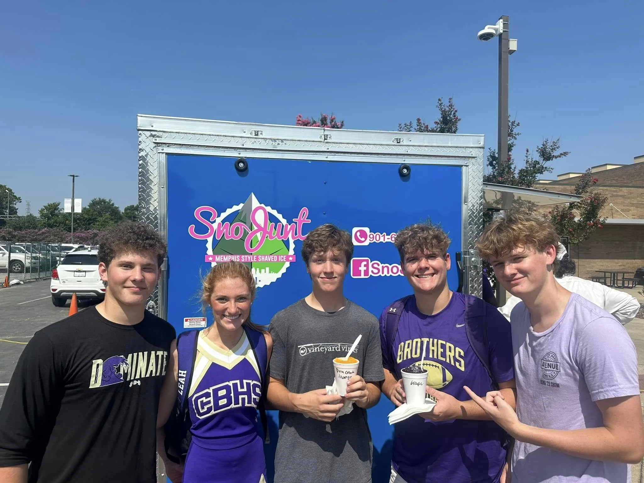 Five teenagers standing in front of a blue Snow Junt ice truck, smiling, holding cups of shaved ice, in a parking lot on a sunny day.