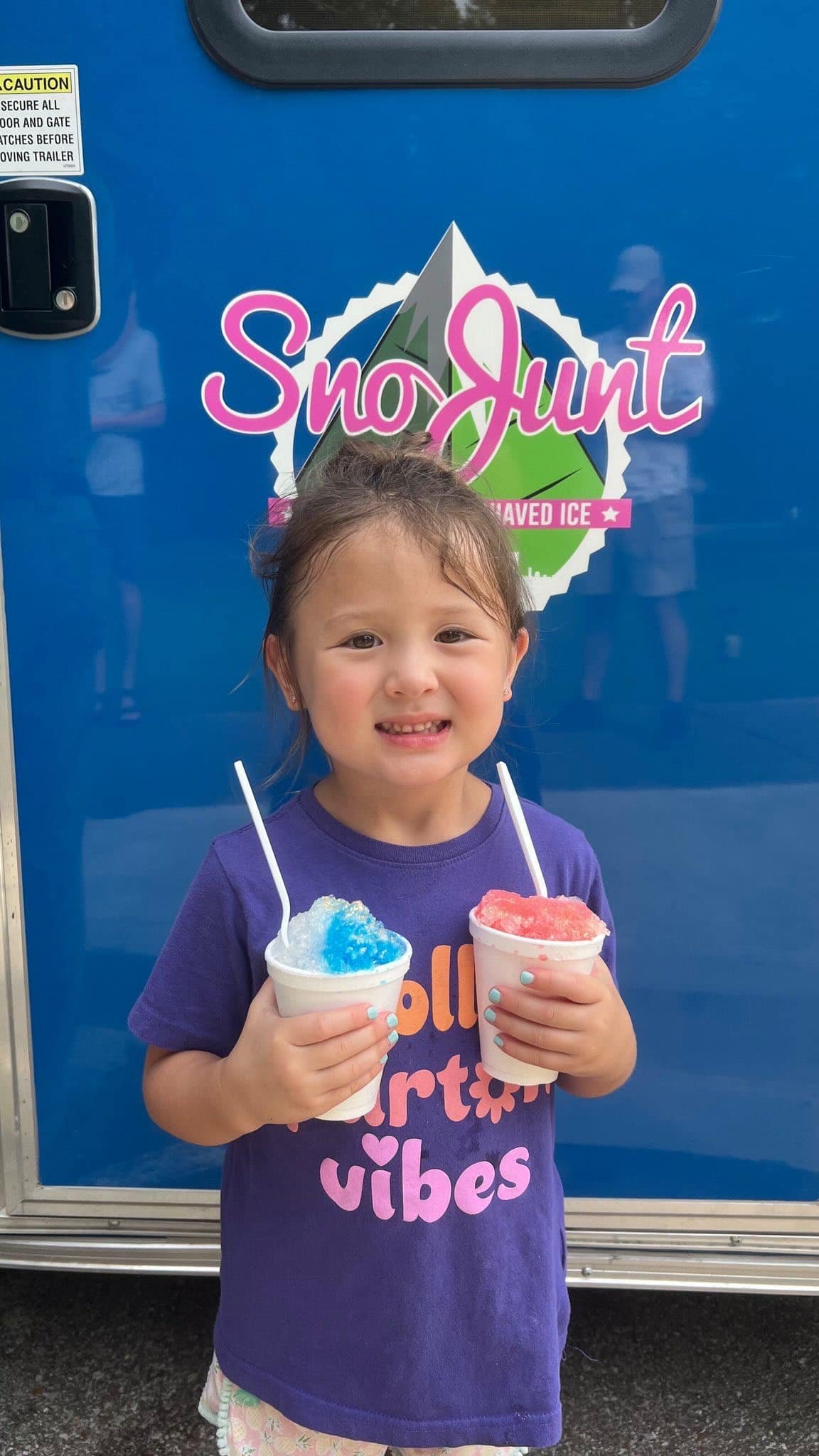 Child holding two shaved ice treats in front of SnoJunt sign on a blue background.