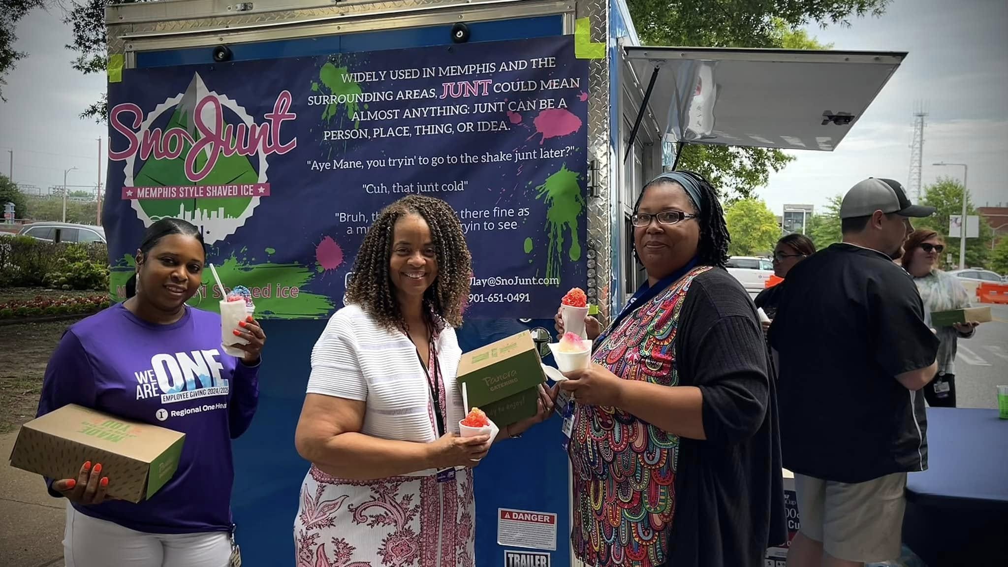 Three women holding shaved ice in front of a food truck with a "Sno Junt" sign and background text. People are gathered around the truck.
