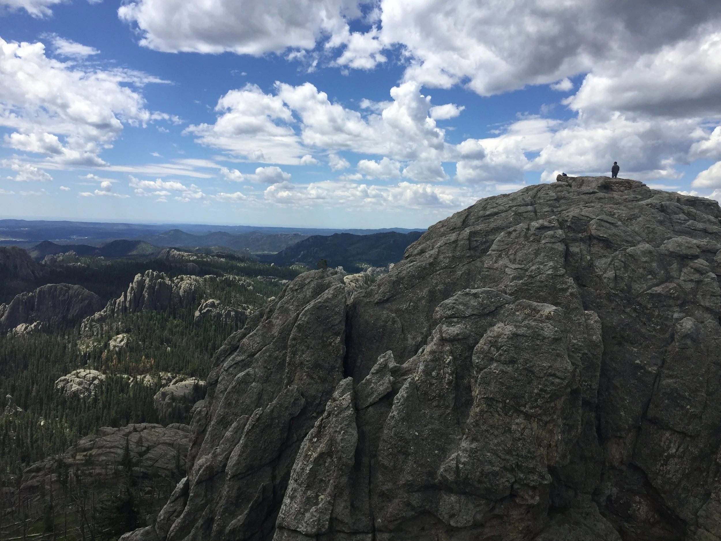 A person standing on top of large rocky mountain overlooking a vast landscape of forests and mountains under a partly cloudy sky.