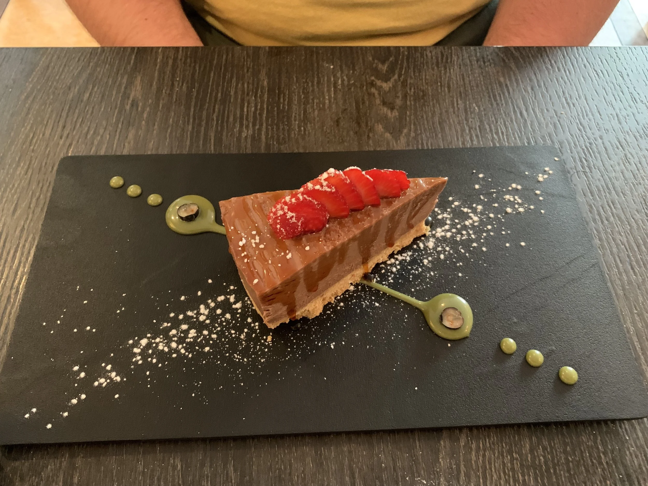 A slice of chocolate cake with strawberries on top, served on a black plate with green sauce droplets and a sprinkle of powdered sugar.