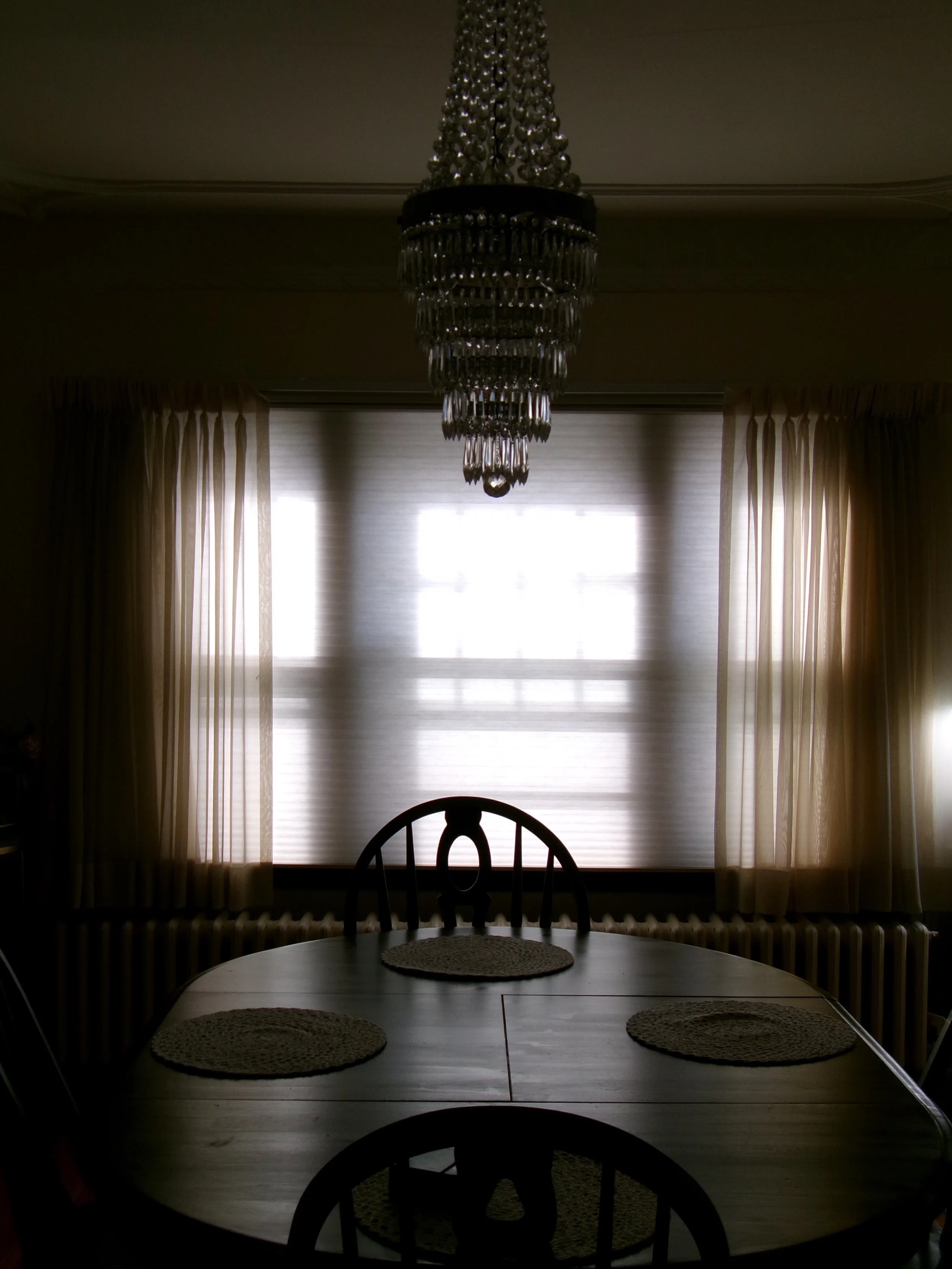 A dining room with a large wooden table, four woven placemats, a high-backed wooden chair, a chandelier hanging from the ceiling, a window with pulled-back curtains, and a radiator beneath the window.