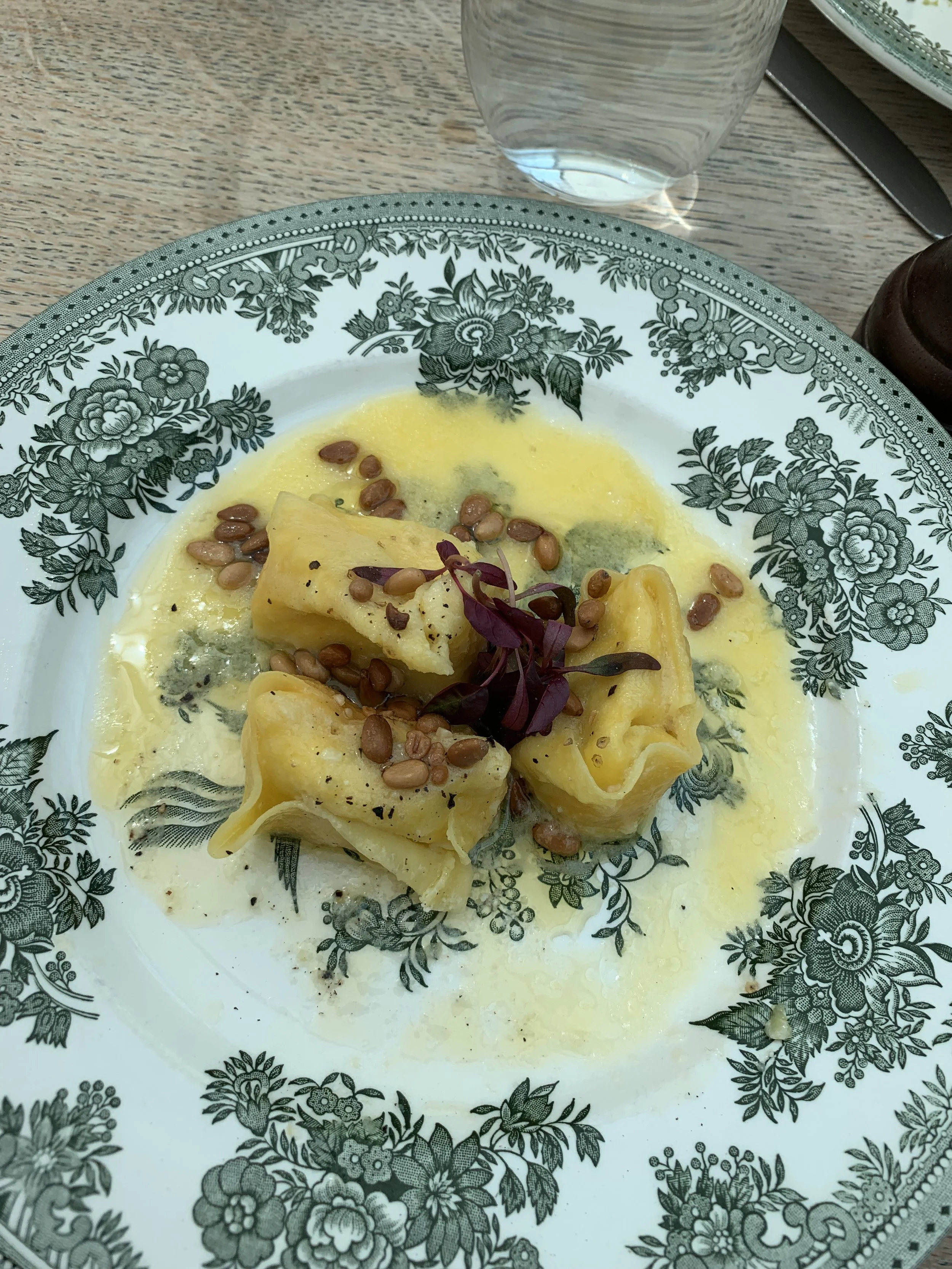 Plate with pasta in a yellow sauce topped with pine nuts and garnished with purple microgreens, set on a floral-patterned plate with a glass of water and black utensils in the background.