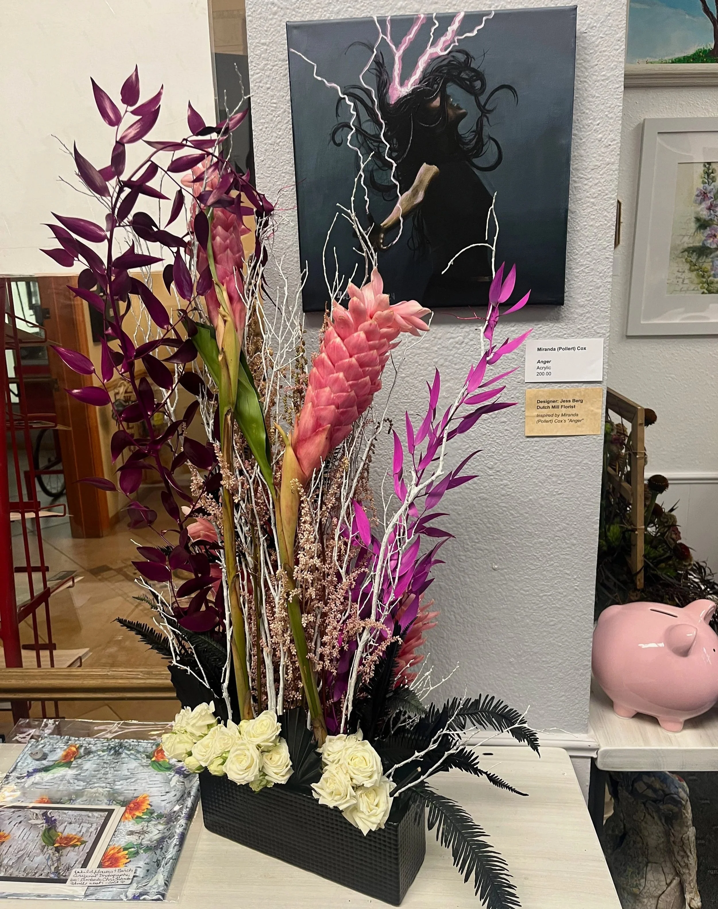 A floral arrangement with white roses, purple leaves, pink ginger flowers, and white branches in a black rectangular vase, placed on a table next to a pink pig-shaped ceramic container.