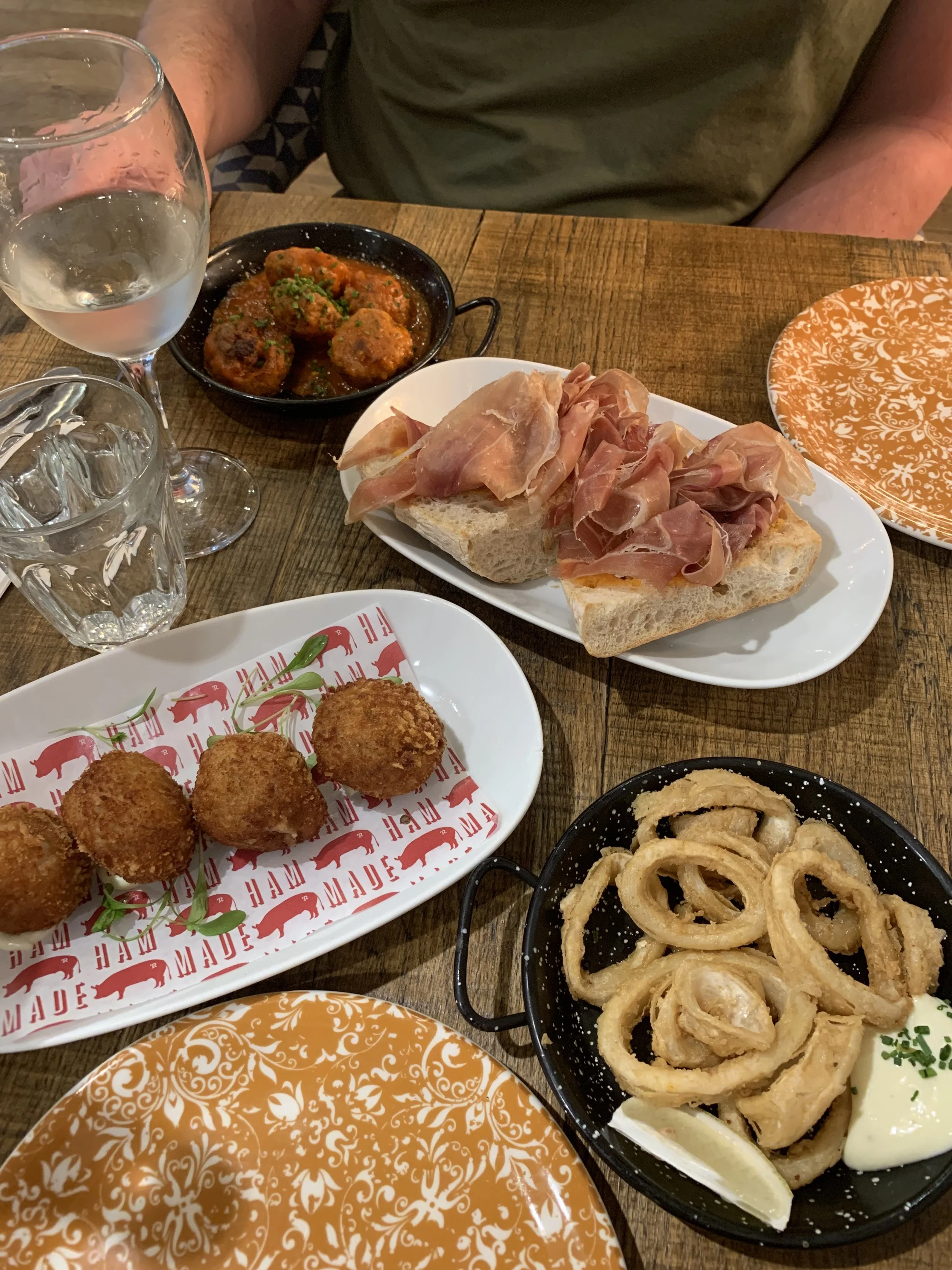 A wooden dining table with various dishes including fried onion rings with dipping sauce, a plate of bread with prosciutto, a small skillet of meatballs in sauce, and a plate of fried croquettes.