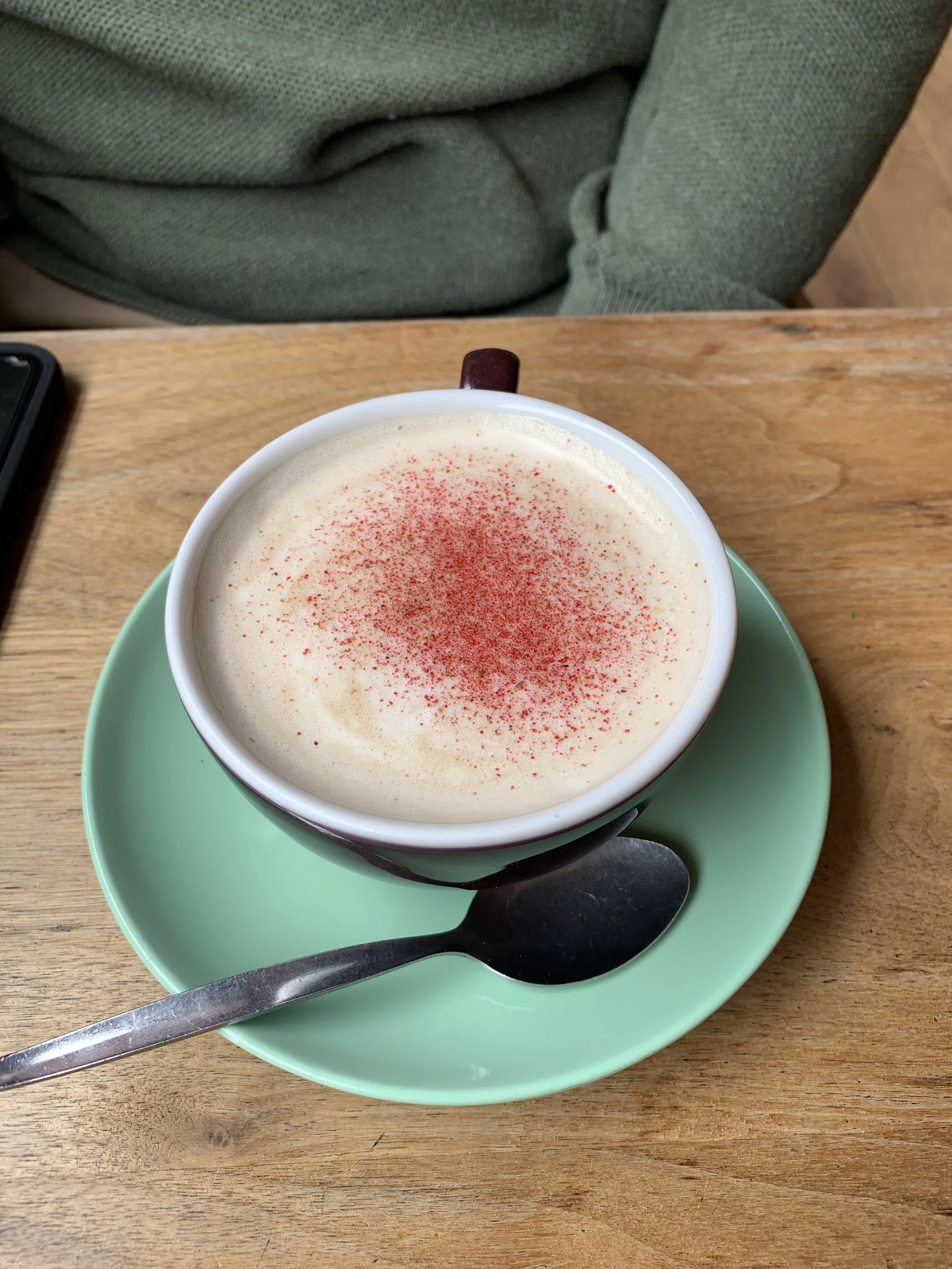 A cup of cappuccino with frothy milk and red sprinkles on top, placed on a mint green saucer with a spoon, on a wooden table.
