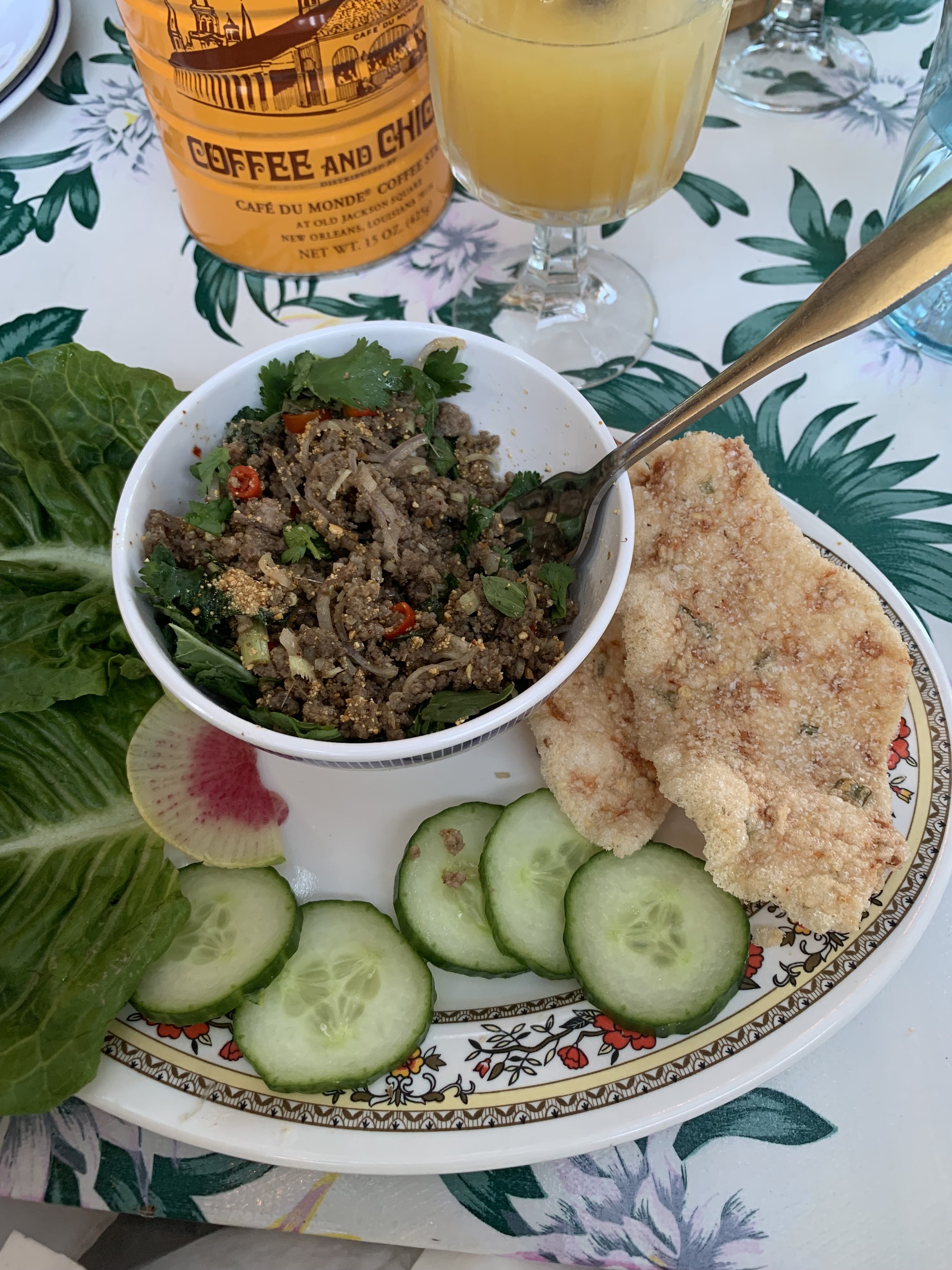 A plate with sliced cucumbers, a fried fish fillet, a bowl of ground meat salad garnished with cilantro and red chili, lettuce leaves, a slice of watermelon radish, and a bottle of coffee with a glass of juice on a floral tablecloth.