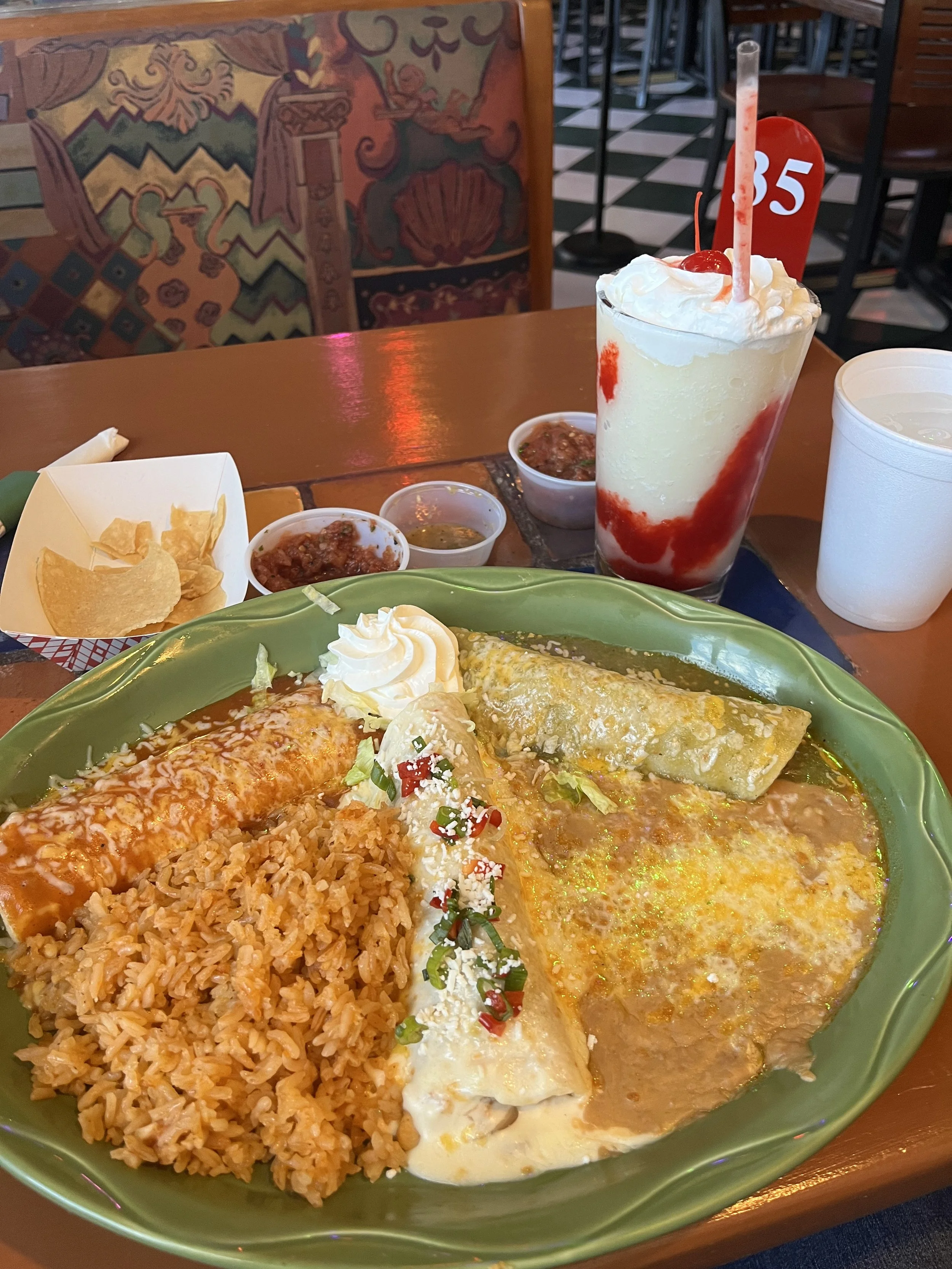 Mexican meal featuring rice, enchiladas, and topped with sour cream and chopped peppers, with a large strawberry milkshake topped with whipped cream and a cherry. There are small bowls of salsa or sauces, a cup of water, and a bag of chips on the tab