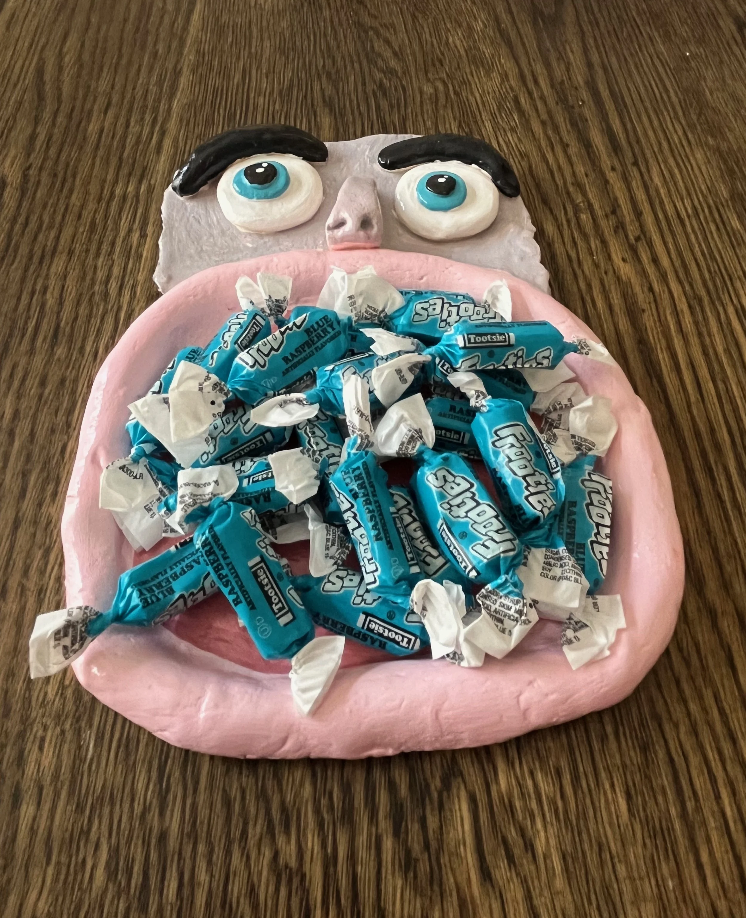 A cake decorated to look like a face with large eyes and black eyebrows, covered with blue and white imported candies on an oval pink base, placed on a wooden surface.