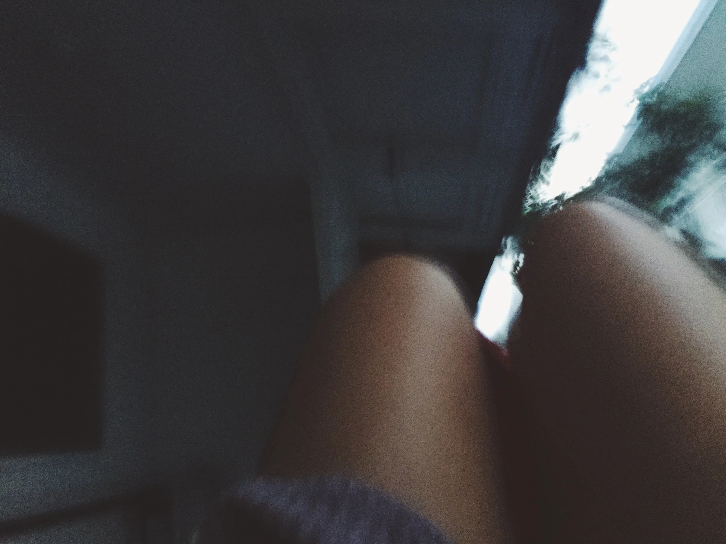 A photograph of a person's legs and knees from a low angle, indoors near a window with some greenery visible outside, possibly sitting on a sofa or similar furniture.