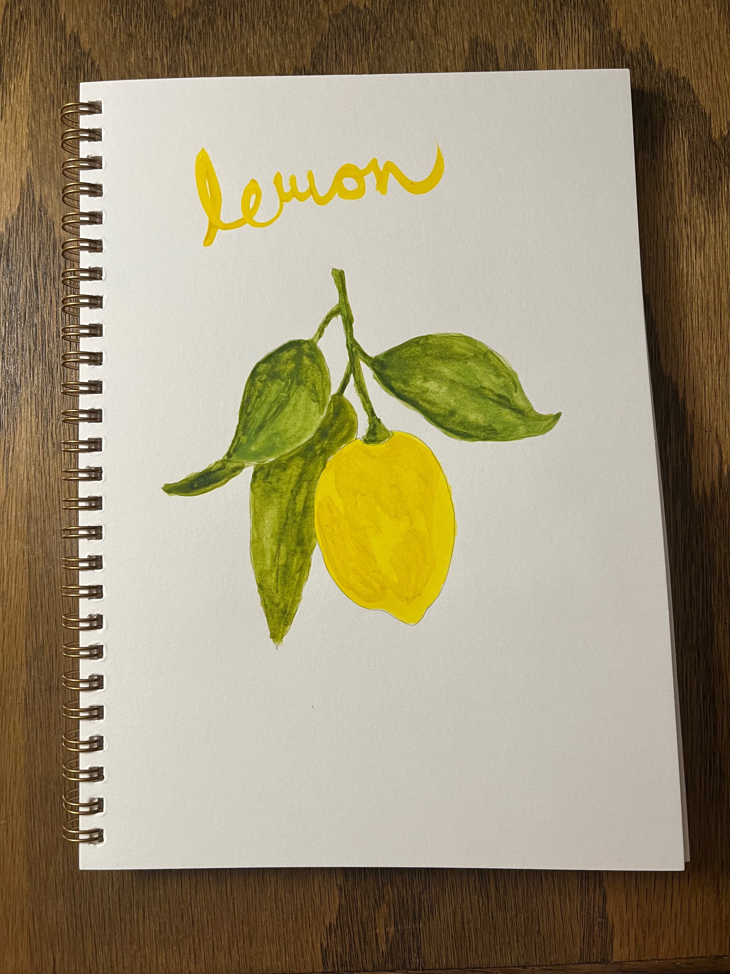 A hand-painted illustration of a lemon with green leaves on a white spiral-bound notebook, placed on a wooden surface. The word 'lemon' is written in yellow cursive at the top.