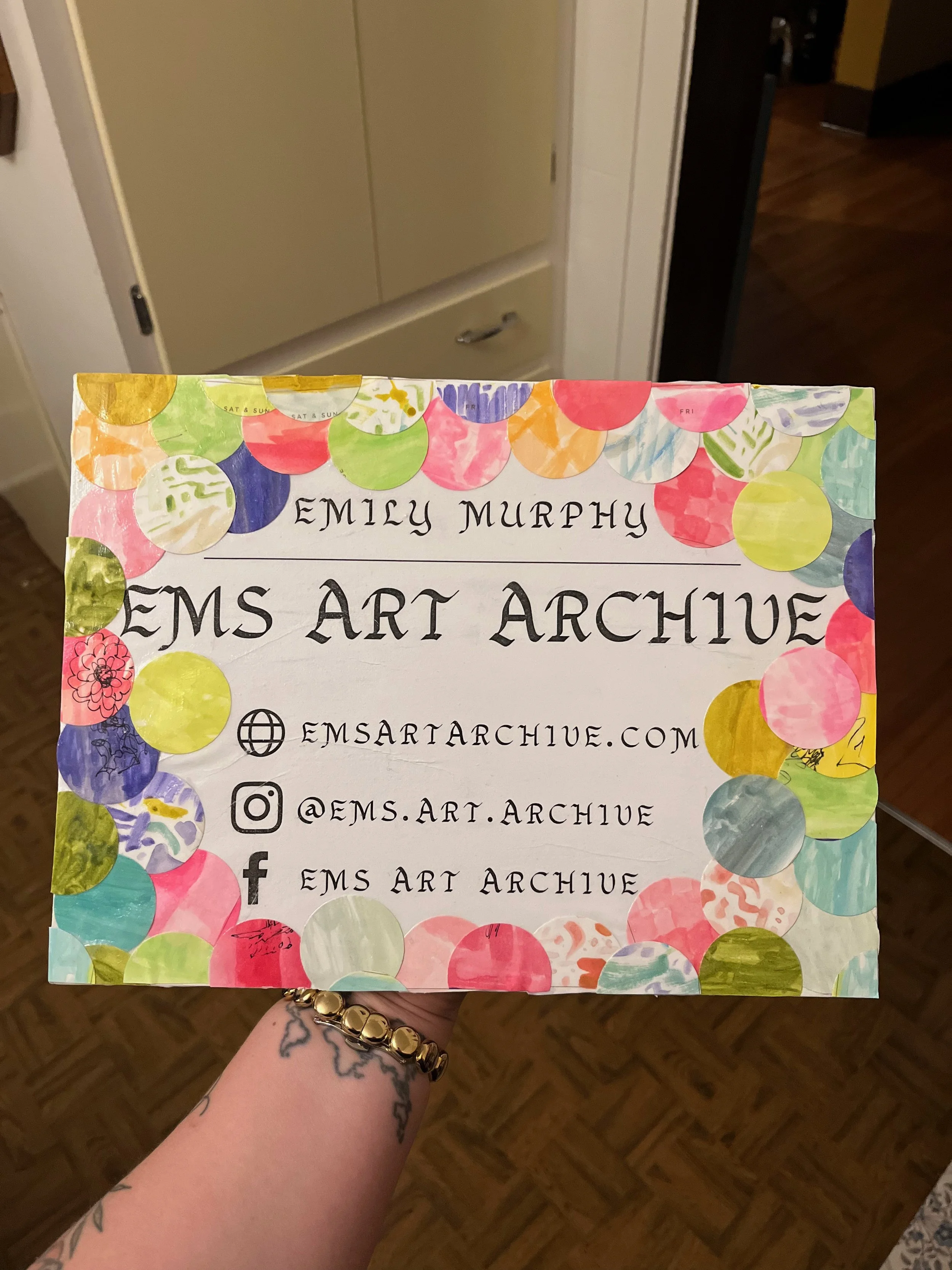 Hand holding an artist's business card with colorful polka dots border, displaying Emily Murphy, EMS Art Archive website, Instagram handle, and Facebook page.