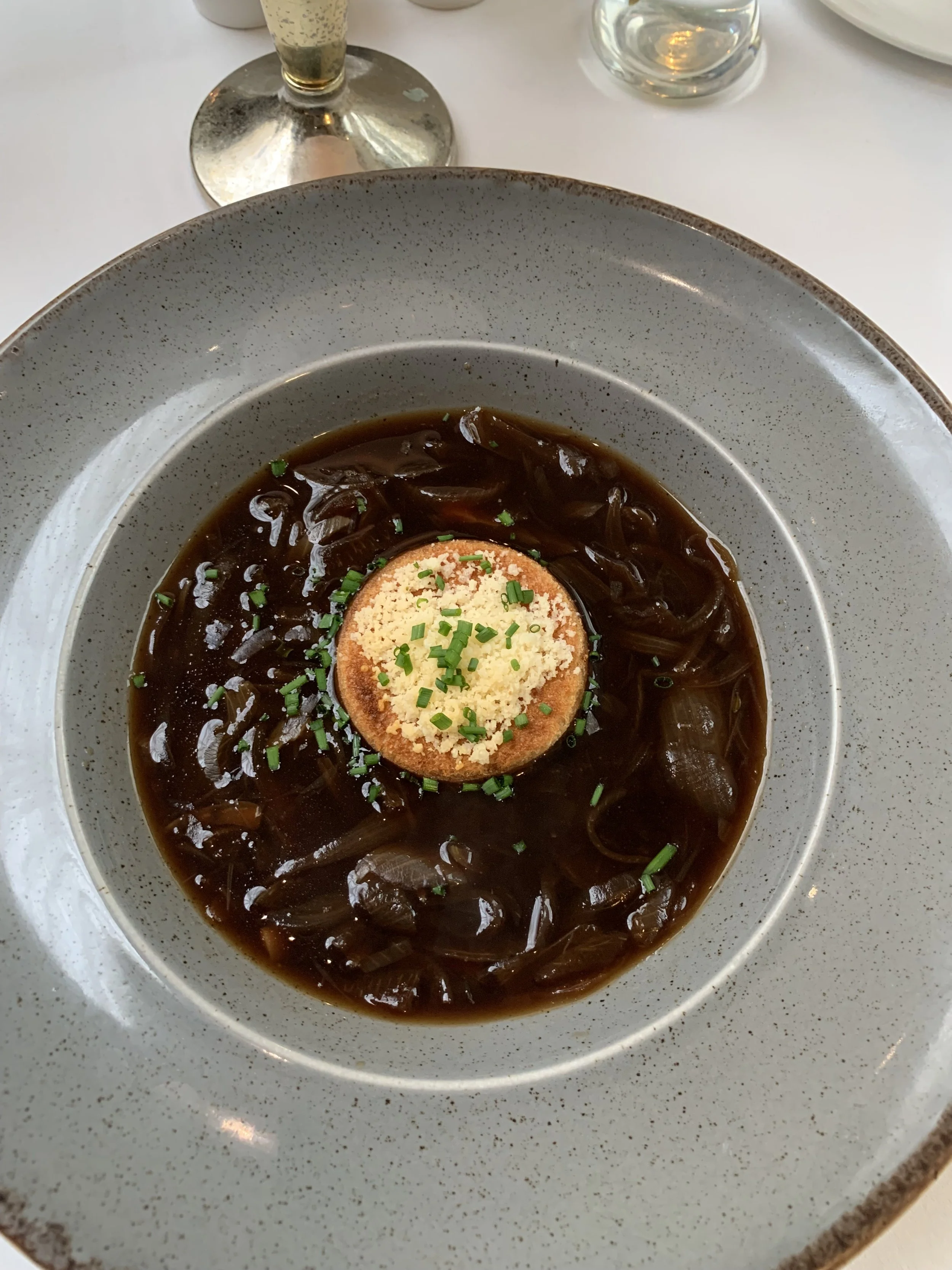 Bowl of French onion soup topped with a toasted bread round, grated cheese, and chopped chives, served on a gray speckled plate.