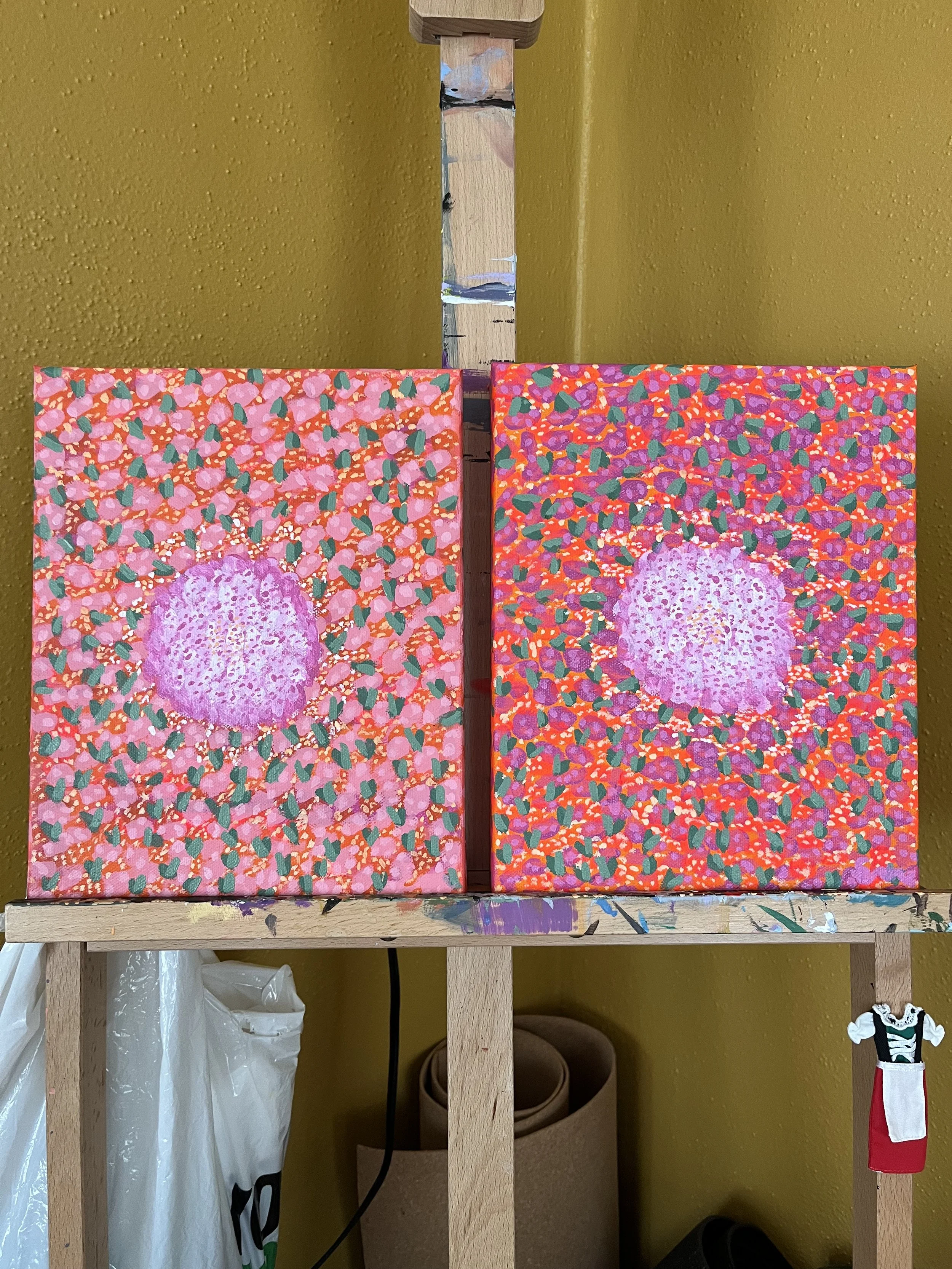 Two colorful abstract paintings on small canvases are displayed on an easel. Each painting features a pinkish-purple central shape surrounded by vibrant, densely packed patterned background with various colors such as pink, green, red, and purple.