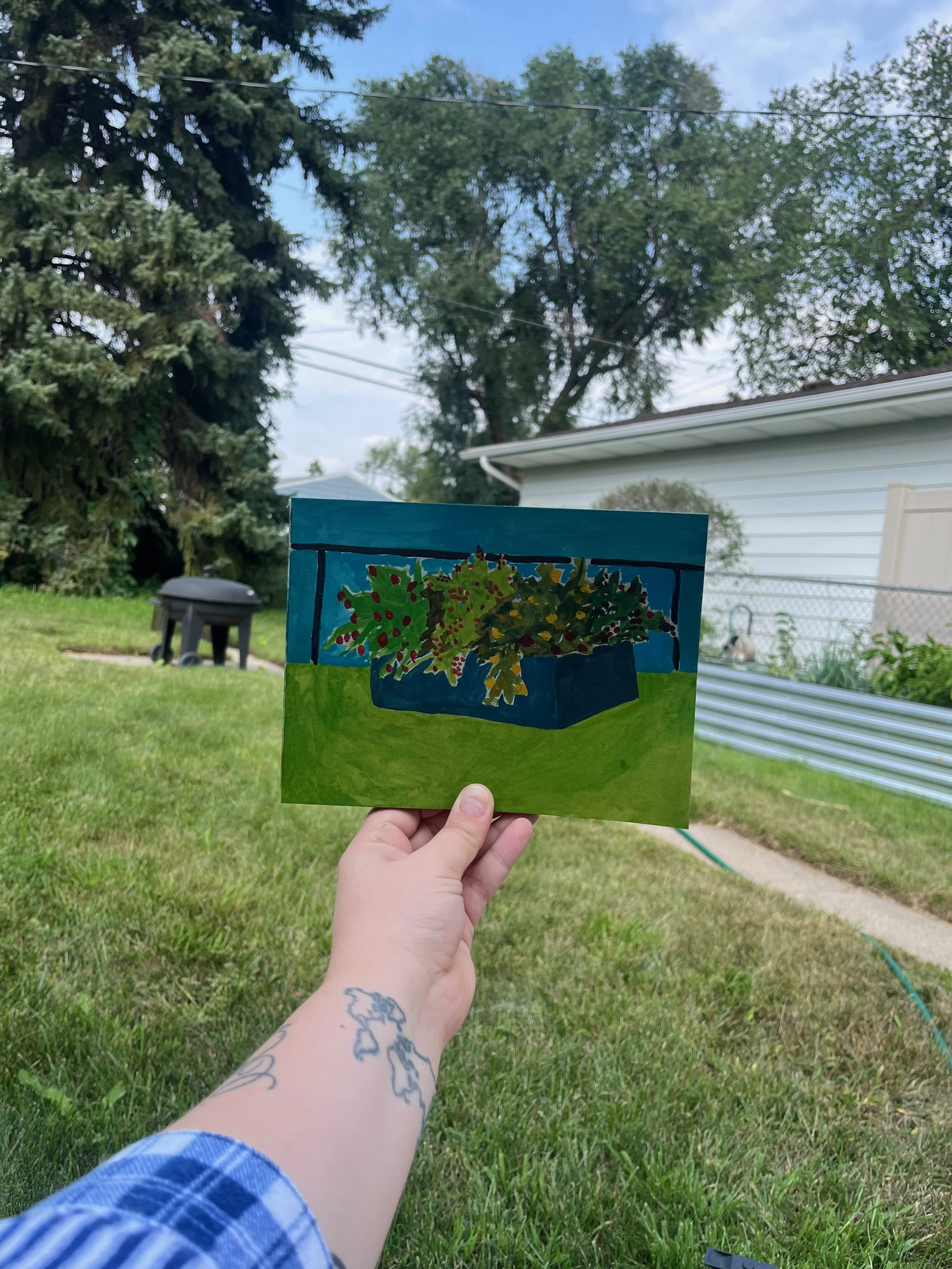 A hand holds a painted artwork of a box of holiday decorations outdoors on a grassy yard with trees and a house in the background.